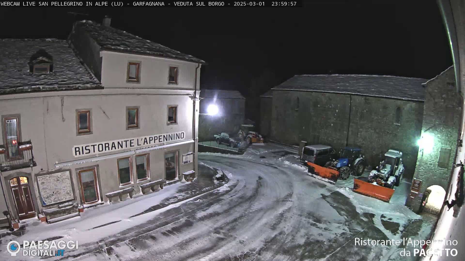 A snow-covered town square at night, illuminated by streetlights, shows a restaurant and several snowplows parked near stone buildings.