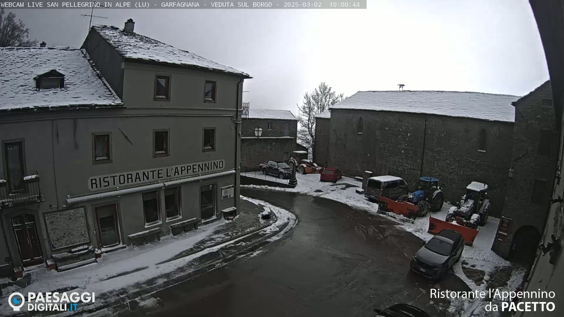 A snow-covered village square with several buildings, vehicles, and snow plows.