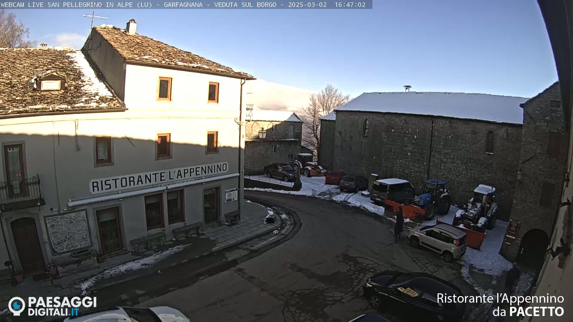 A sunny, snow-dusted village square with several parked cars and tractors near stone buildings, one of which is a restaurant.