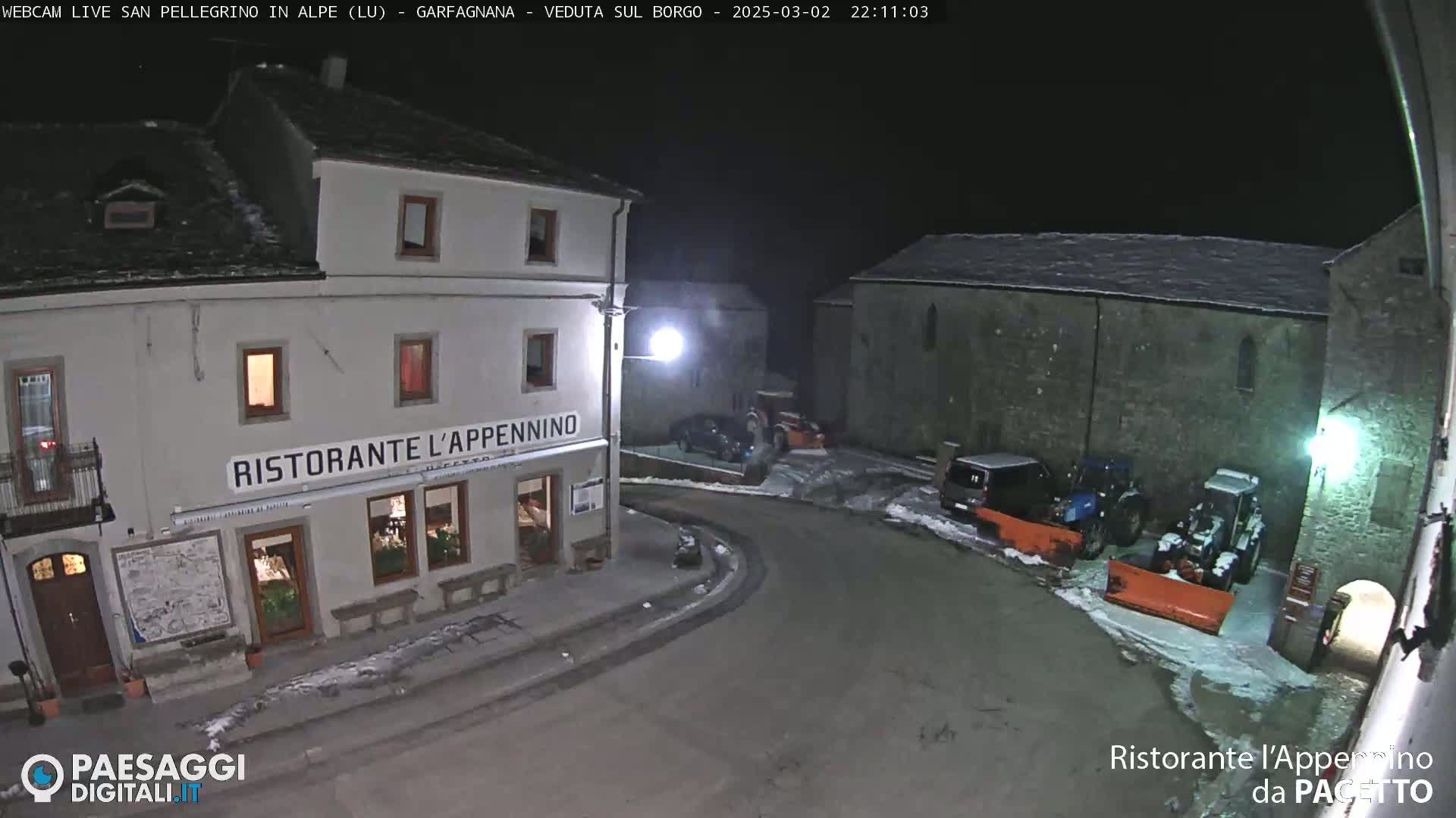 A snow-covered nighttime village square features a restaurant, several parked vehicles, and snowplows.