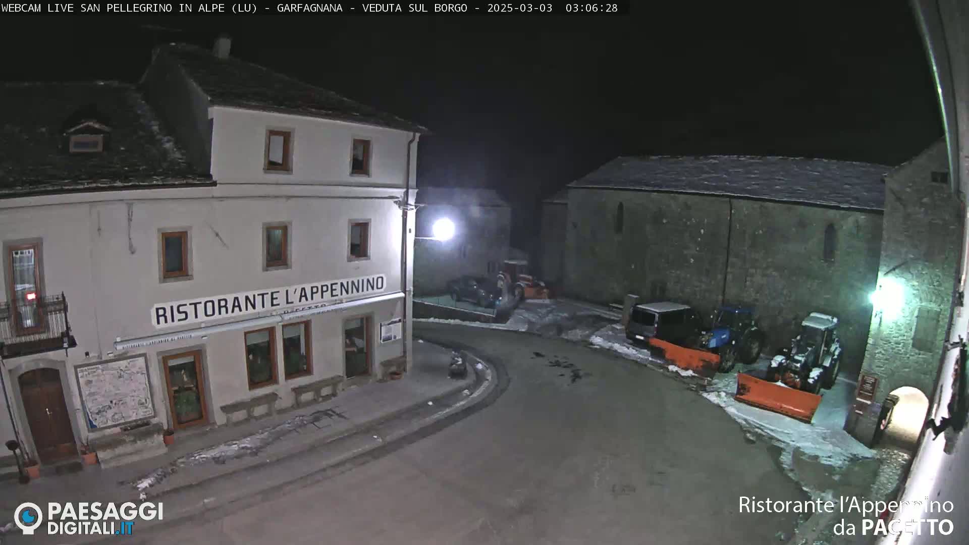 A snow-covered village square at night, illuminated by streetlights, with several parked vehicles near a restaurant and snowplows.