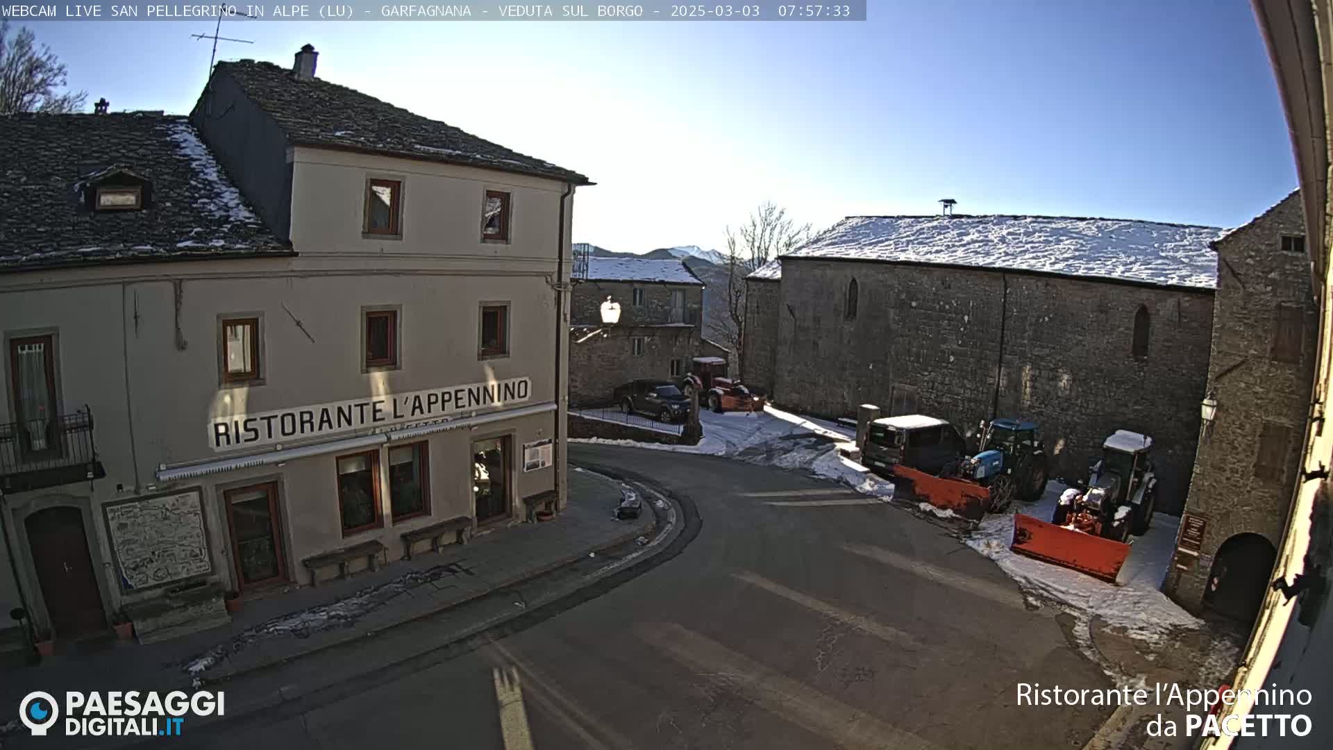 A sunny, snow-dusted village square with several buildings, a few vehicles, and snow plows.