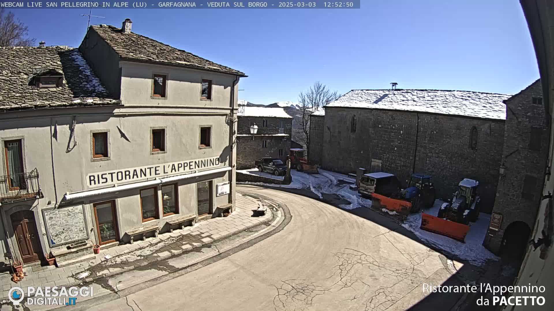 A sunny day in a snow-dusted village square shows several buildings, some vehicles, and snow removal equipment.