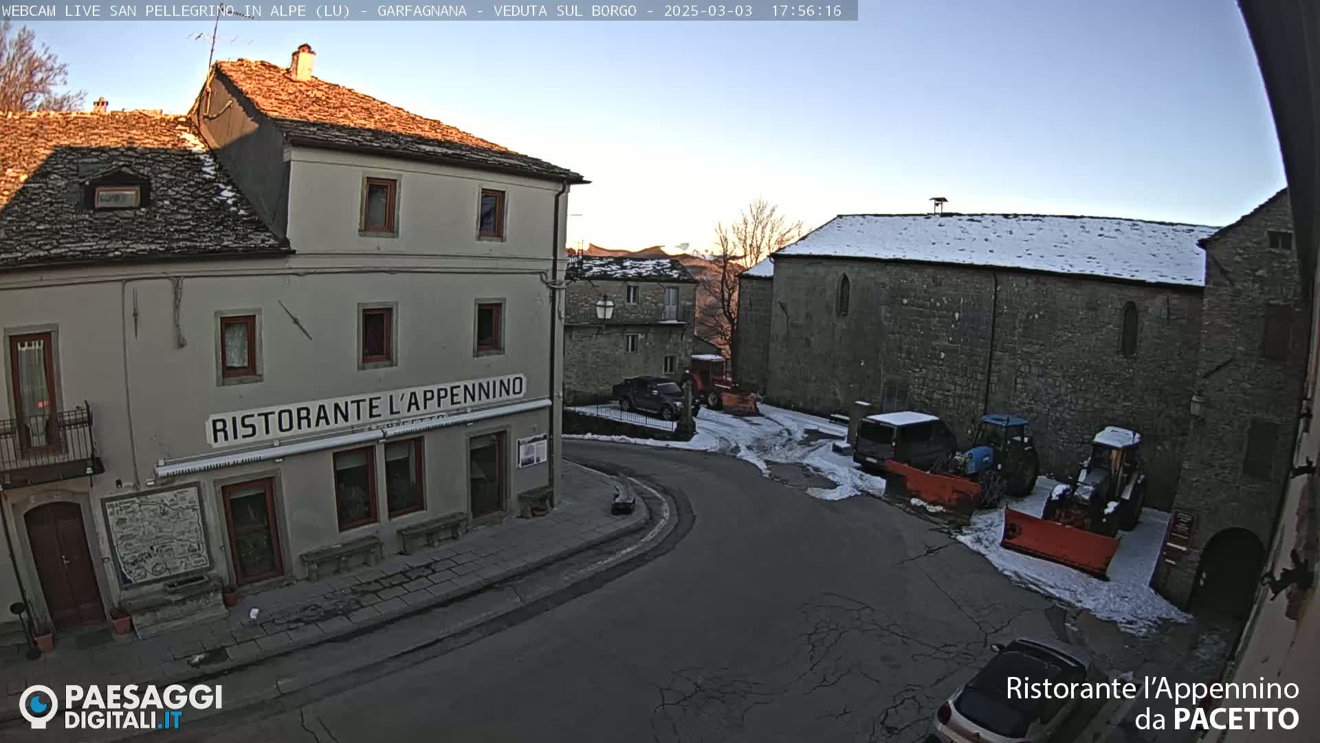 A quaint village square with stone buildings, some snow on the ground, and several parked vehicles, including snow removal equipment, under a clear sky.