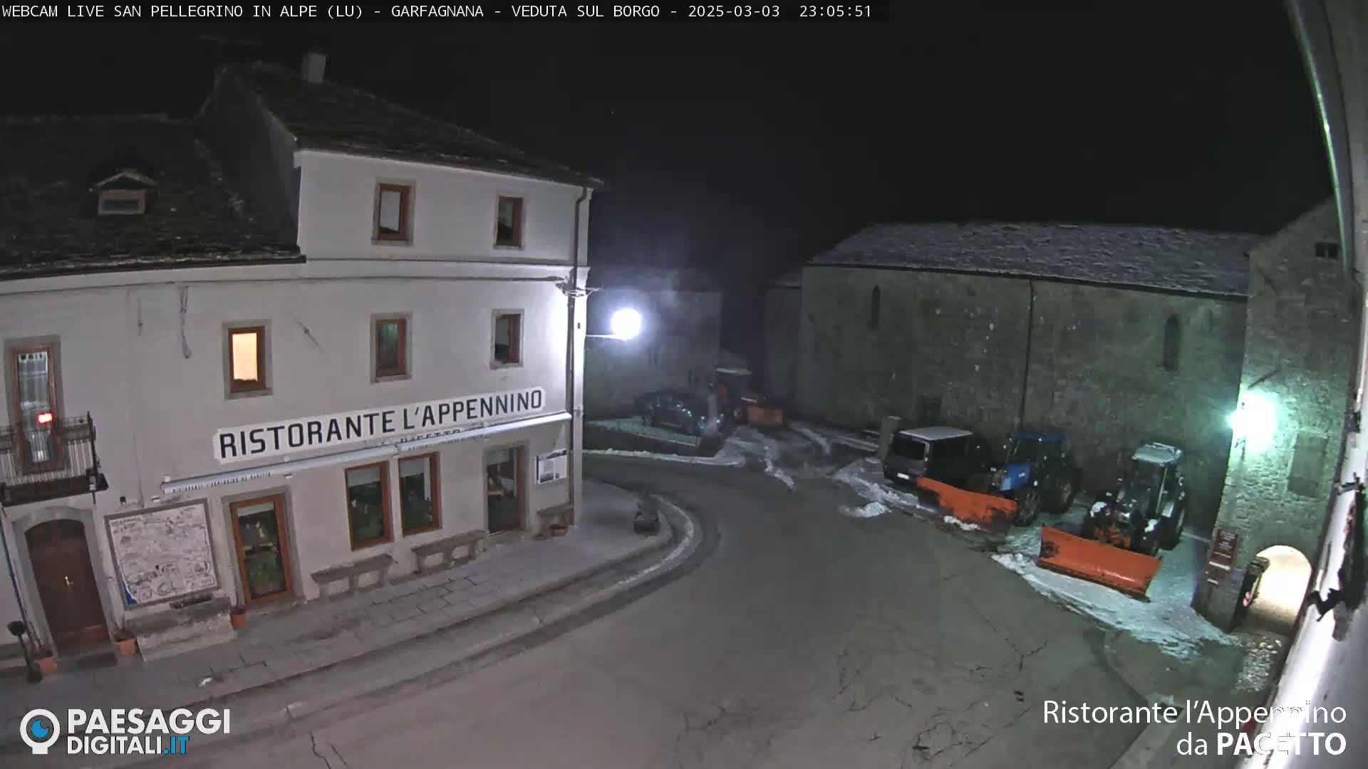 A nighttime view of a snow-dusted village square with a restaurant, parked vehicles, and snowplows.