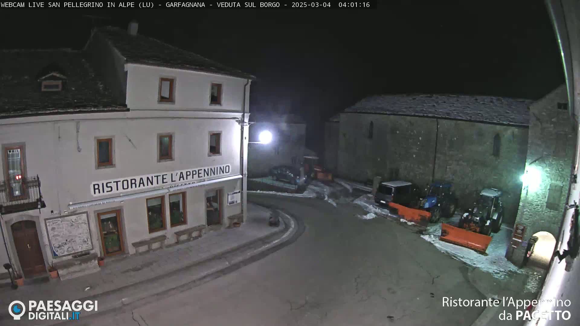 A snow-covered town square at night is illuminated, showing a restaurant and parked vehicles, including snowplows.