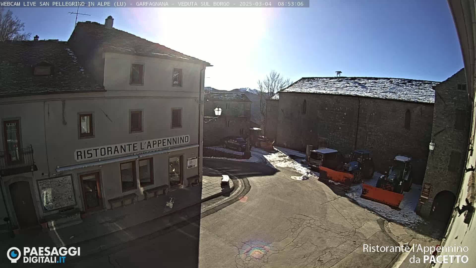 A sunny, snow-dusted village square with several buildings, some parked vehicles, and snow plows.
