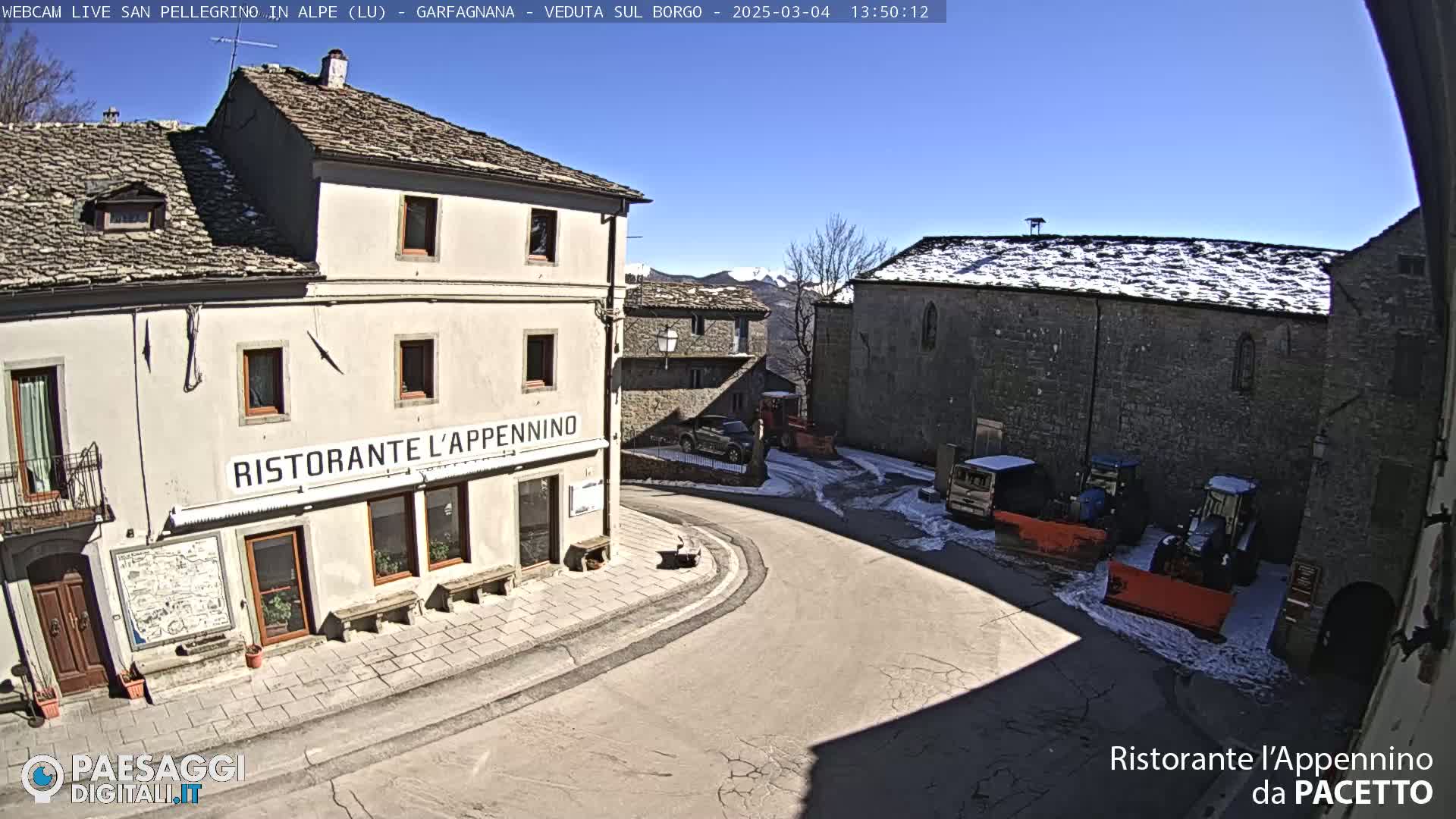 A sunny day in a stone village square with snow on some rooftops, where several vehicles, including snowplows, are parked near a restaurant.