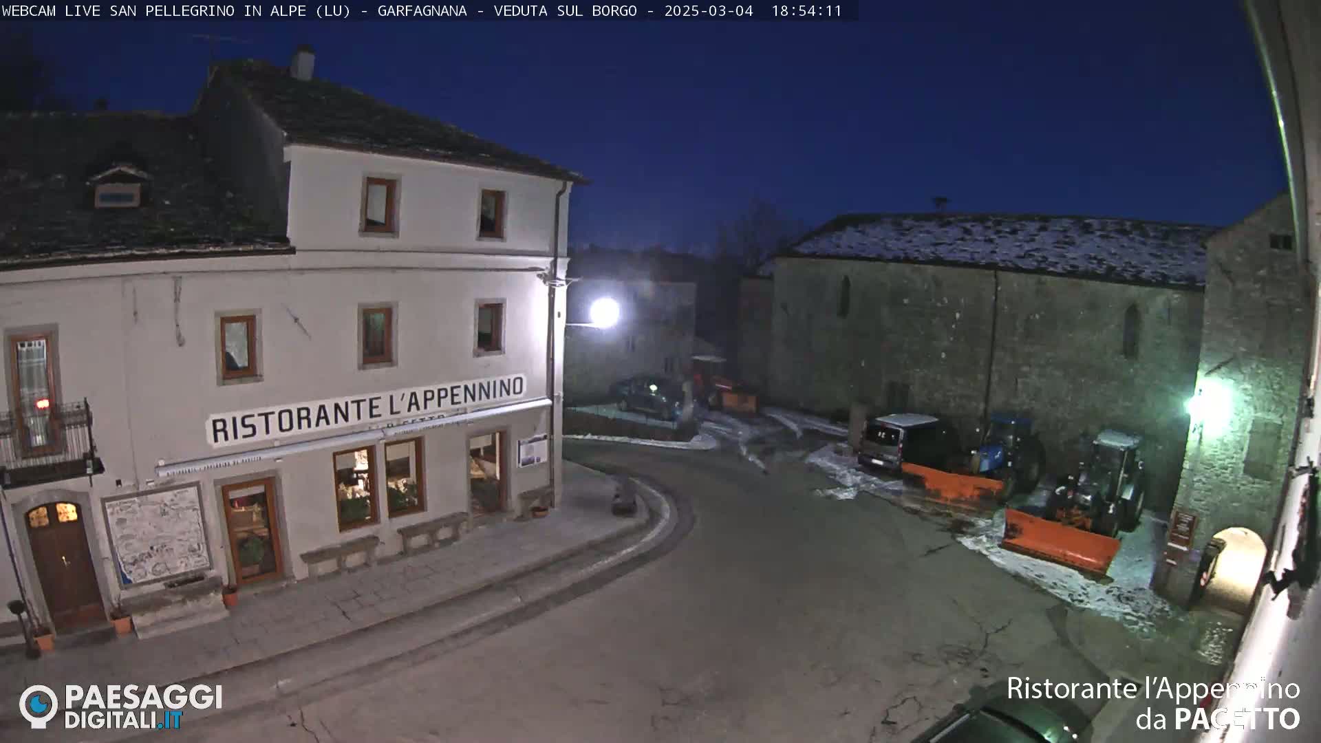 A nighttime view of a town square with snow on the ground, featuring a restaurant called Ristorante l'Appennino and snow-clearing machinery.