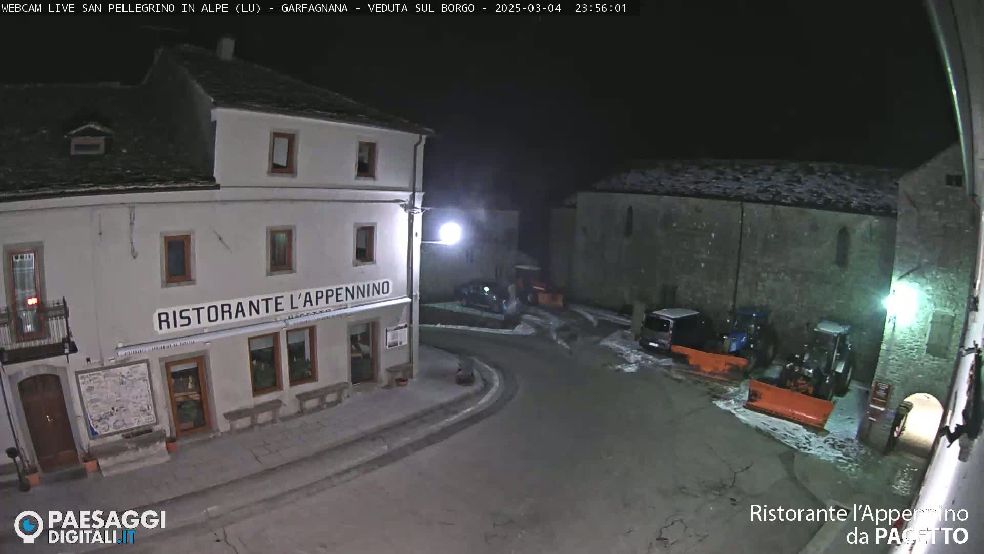 A snow-dusted nighttime village square shows a restaurant, parked vehicles, and snow-clearing equipment.