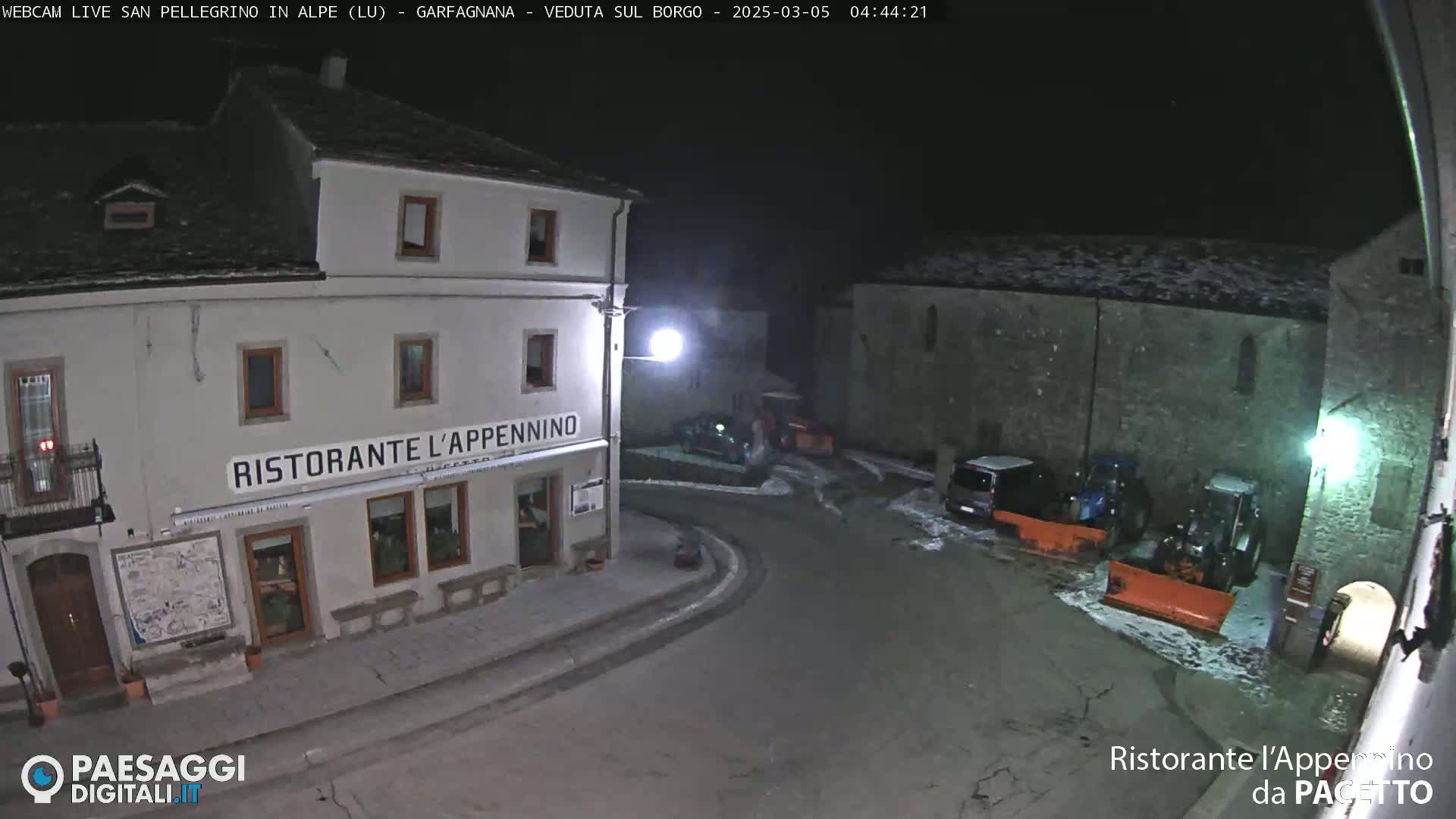 A nighttime, snow-dusted village square features a restaurant and parked vehicles, with snow plows present.