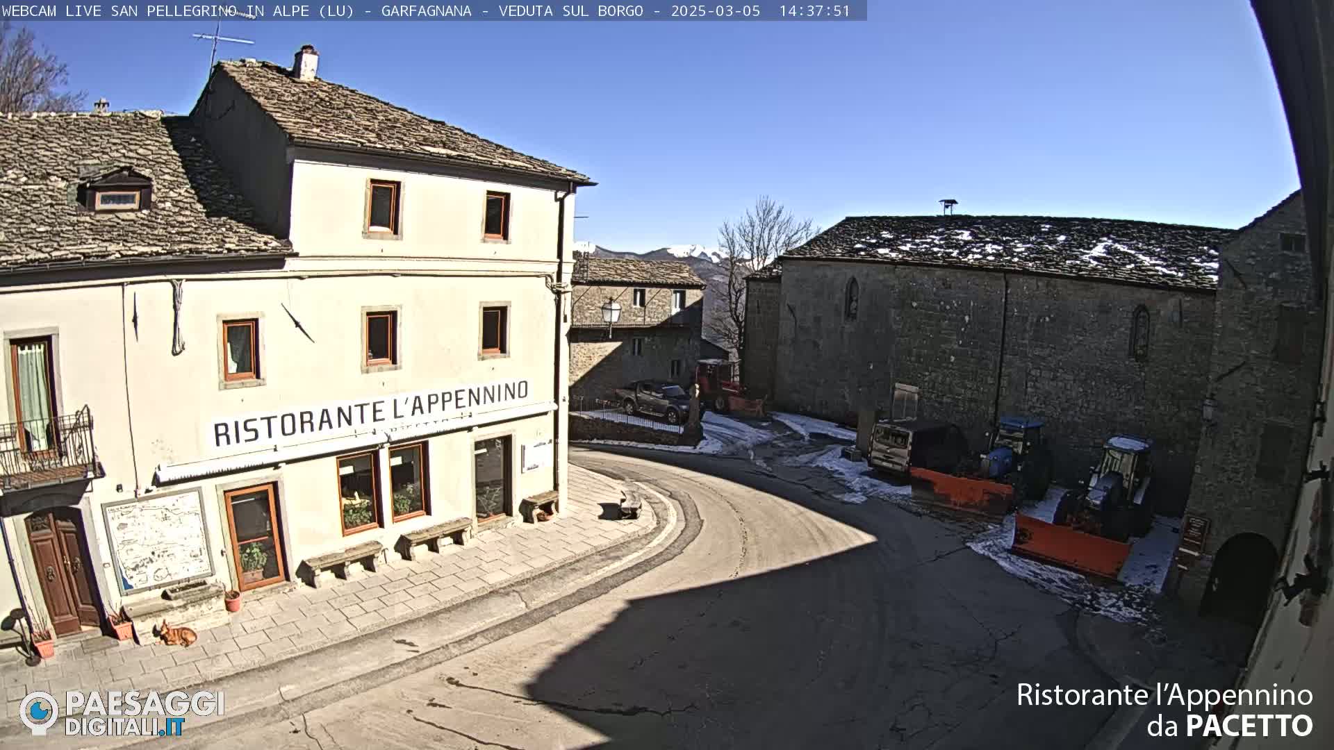 A sunny, snow-dusted village square features a restaurant, several buildings, and some parked vehicles, including snowplows.
