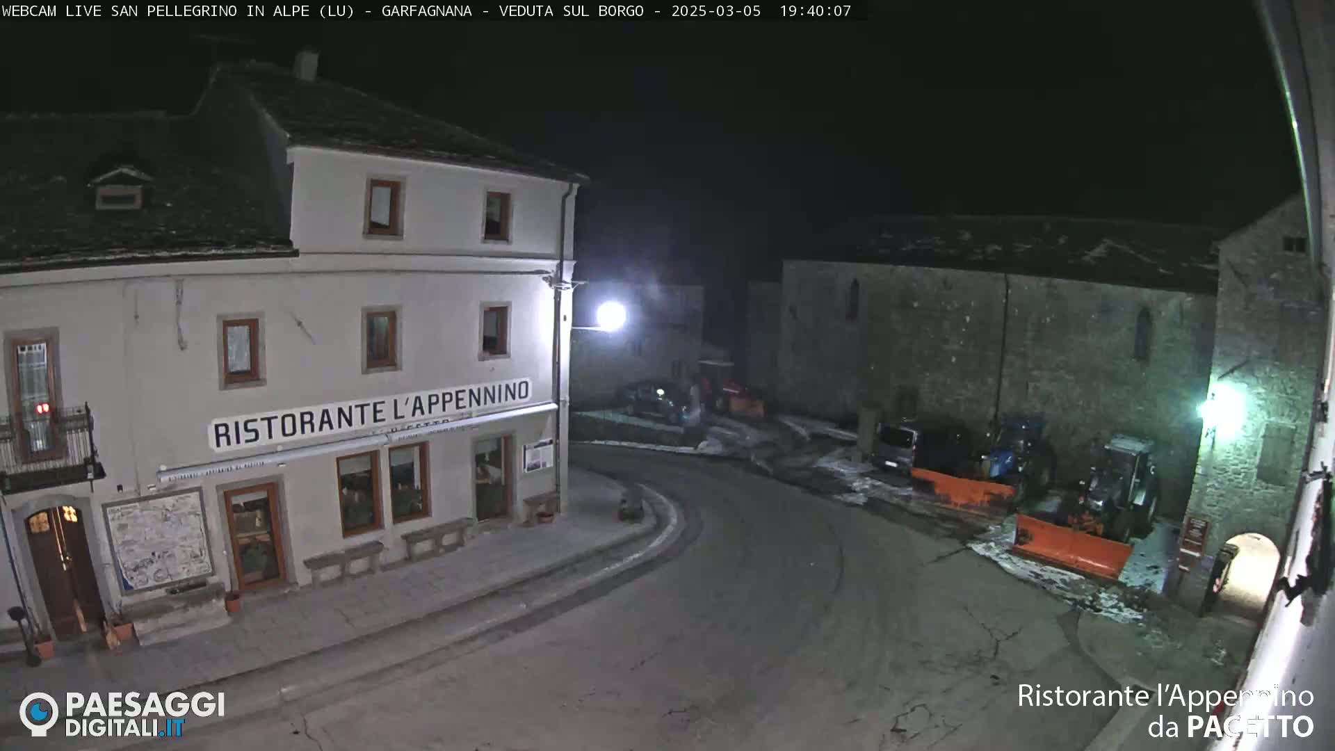 A nighttime, snow-covered village square with a restaurant called "Ristorante L'Appennino," and several parked vehicles, including snowplows.