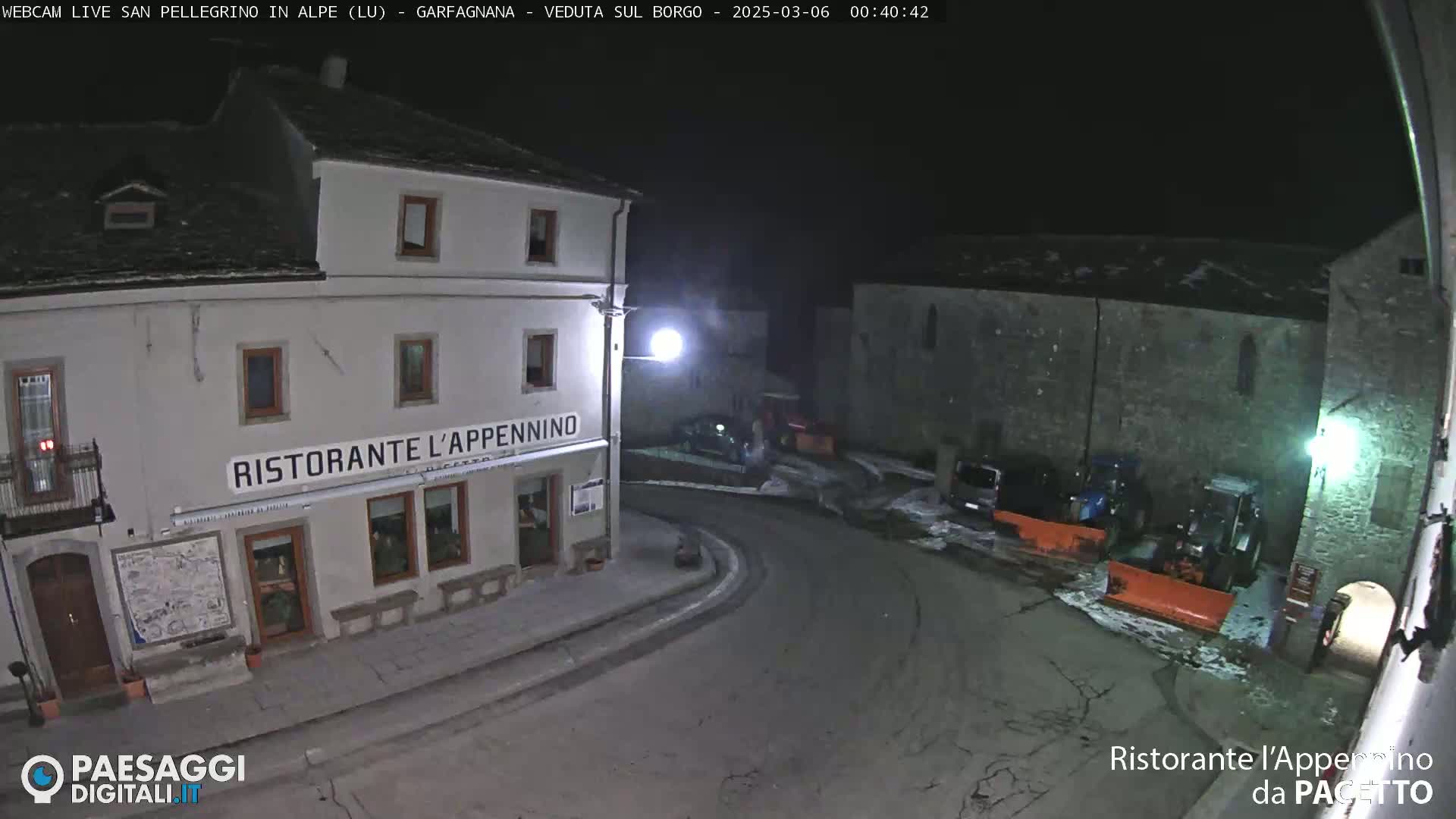 A nighttime, snow-covered village square with a restaurant, some parked cars, and snow-removal equipment.