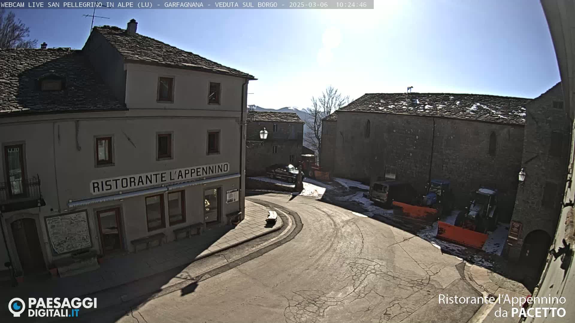 A sunny day in a quaint stone village square, where a restaurant sits beside a curving road, with snow patches visible and some parked vehicles including snowplows.