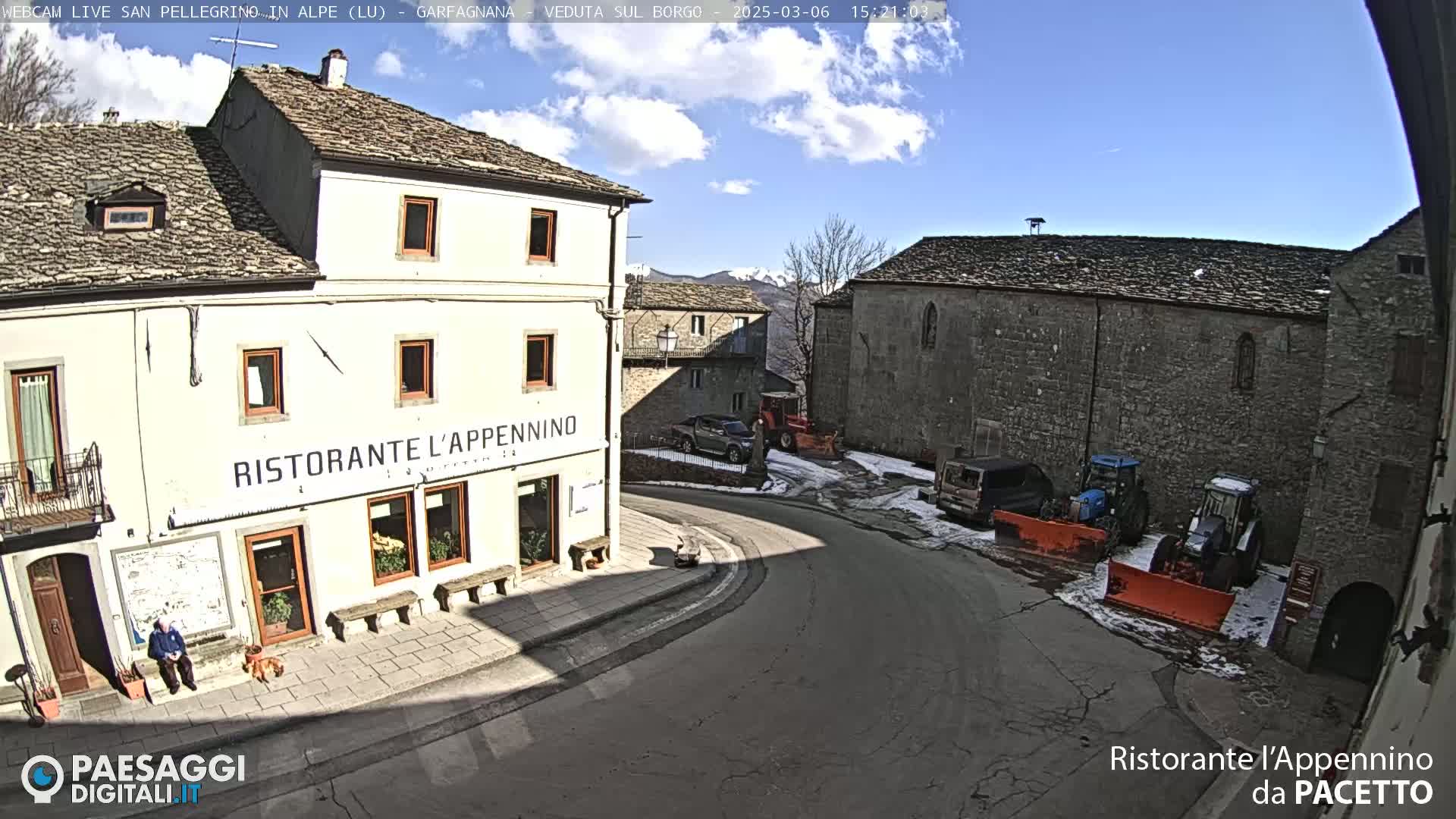 A sunny day reveals a quaint village square with stone buildings, a restaurant, parked vehicles, and snow removal equipment.