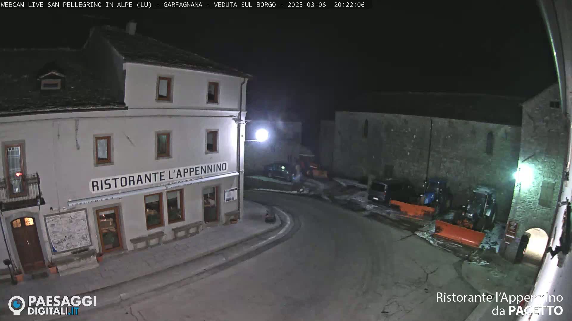 A nighttime view of a snow-covered village square with a restaurant, some parked cars, and snow removal equipment.