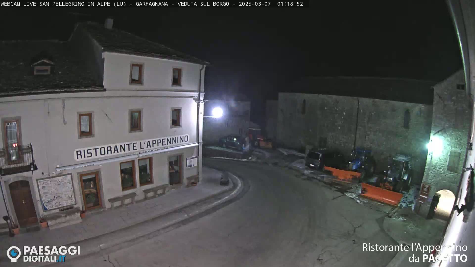 A nighttime view of a town square with snow on the ground, featuring a restaurant called "Ristorante l'Appennino" and snowplows parked nearby.