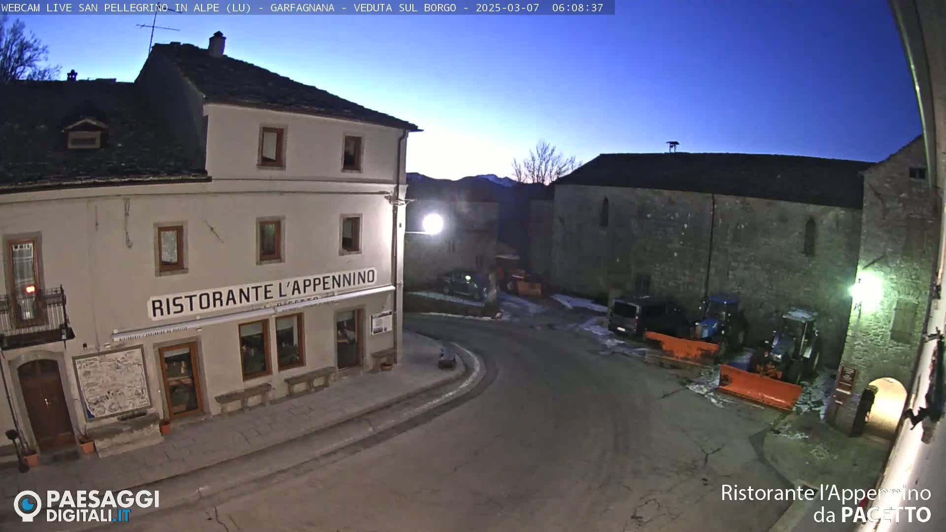 A mostly empty town square at twilight, with a restaurant, stone buildings, and snowplows parked nearby under a clear, dark sky.