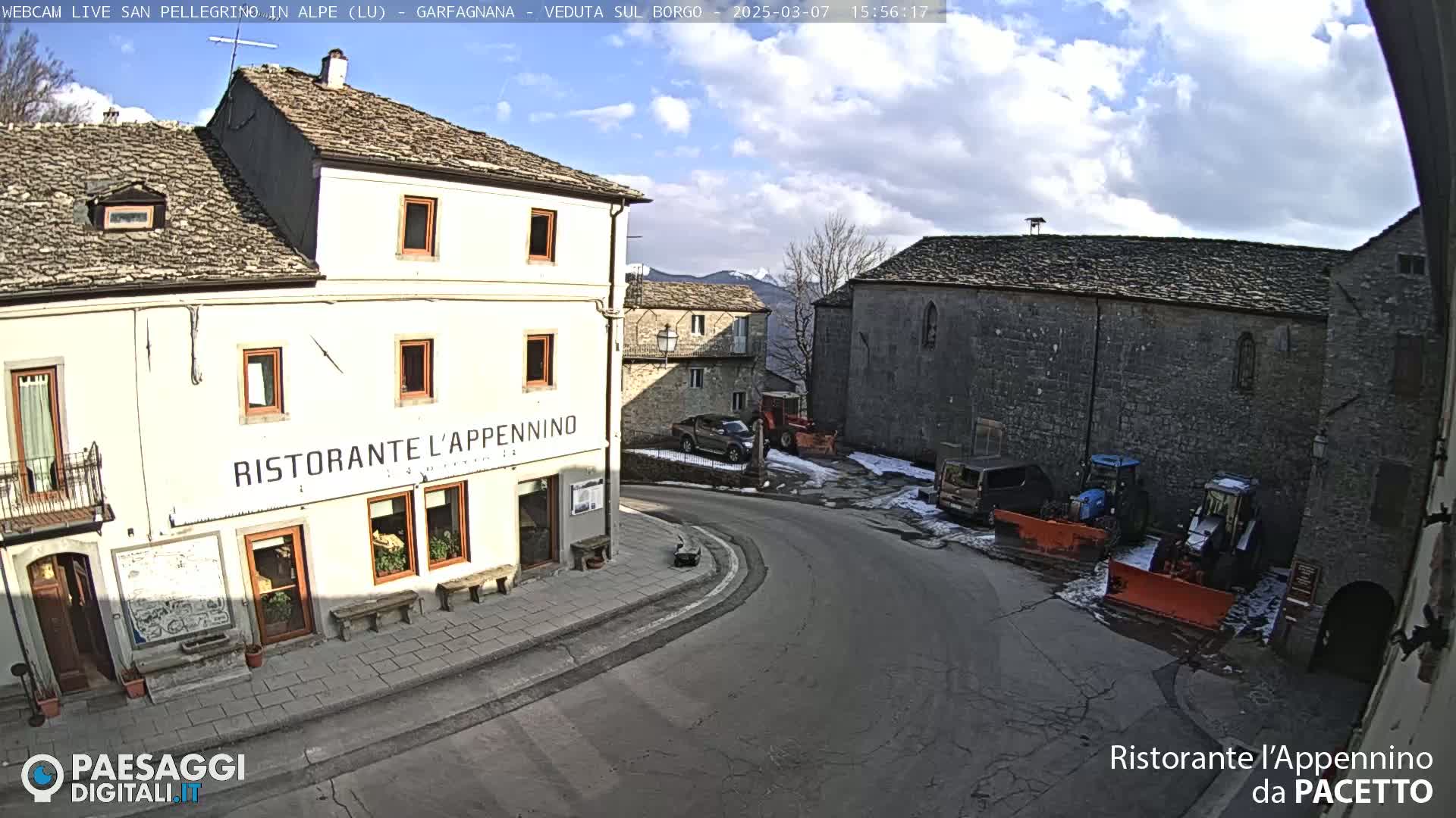 A partly sunny day in a small village square with stone buildings, some vehicles, and snow removal equipment.