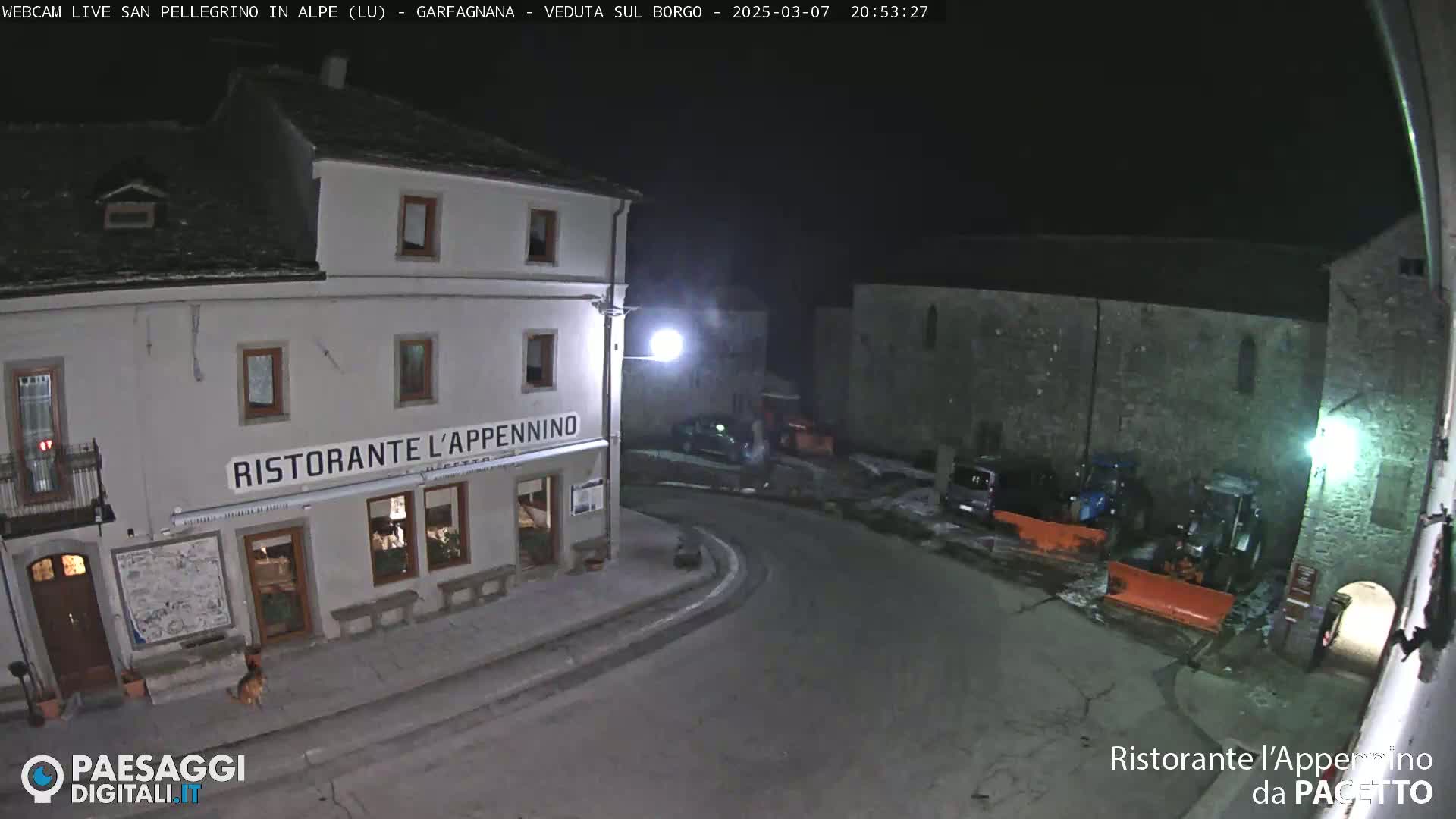 A nighttime view of a town square with a restaurant, some parked vehicles, and snow on the ground.