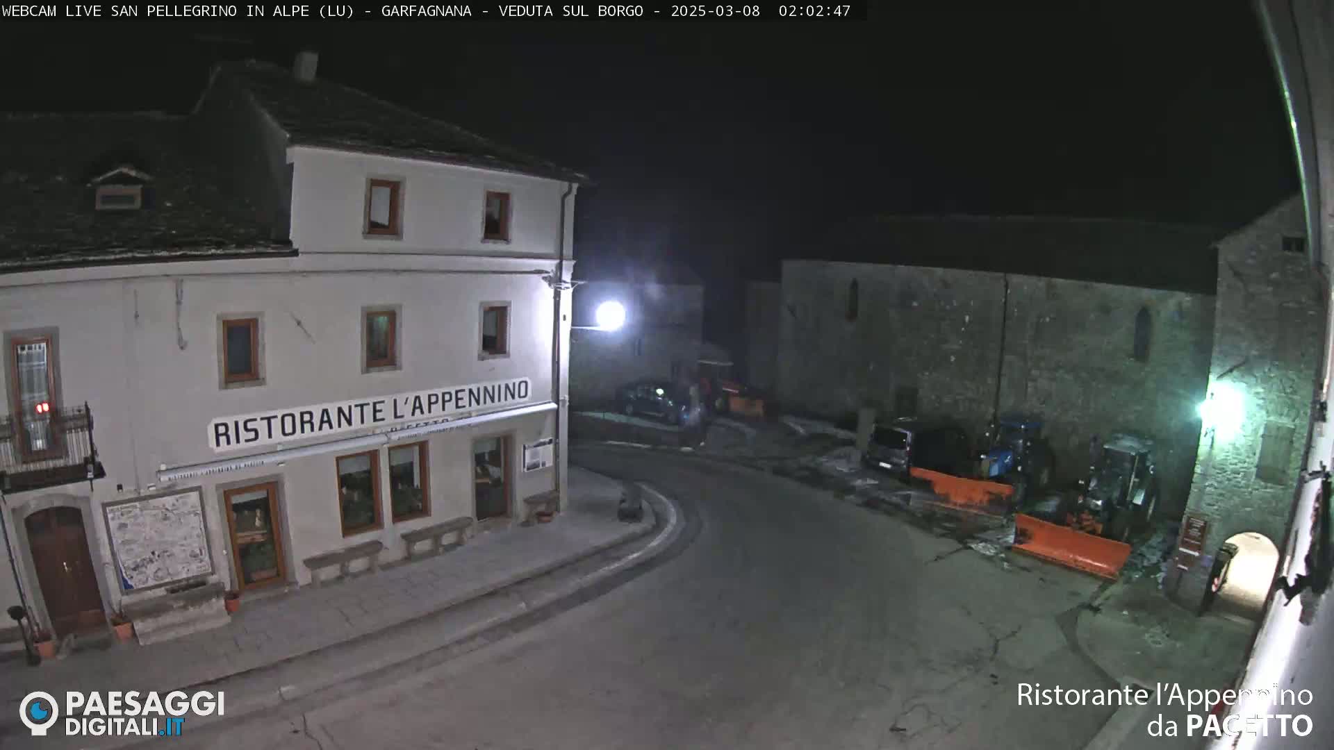 A nighttime view of a town square with a restaurant, some vehicles, and snow-covered ground.