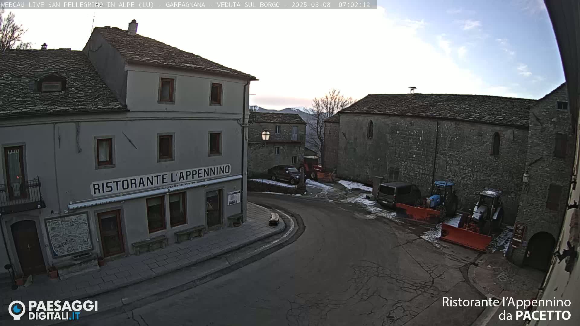 A mostly empty town square with a restaurant, several parked vehicles, and snow removal equipment is visible under a mostly clear sky.
