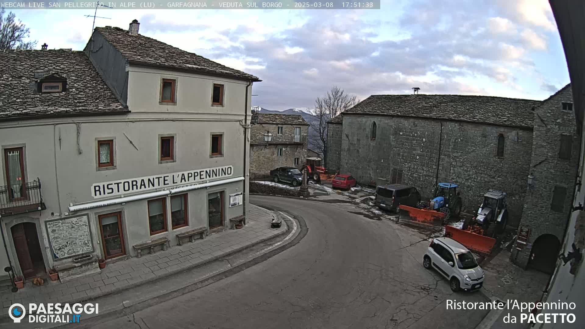 A quaint village square with stone buildings, a restaurant, several vehicles, and snow-removal equipment under a partly cloudy sky.