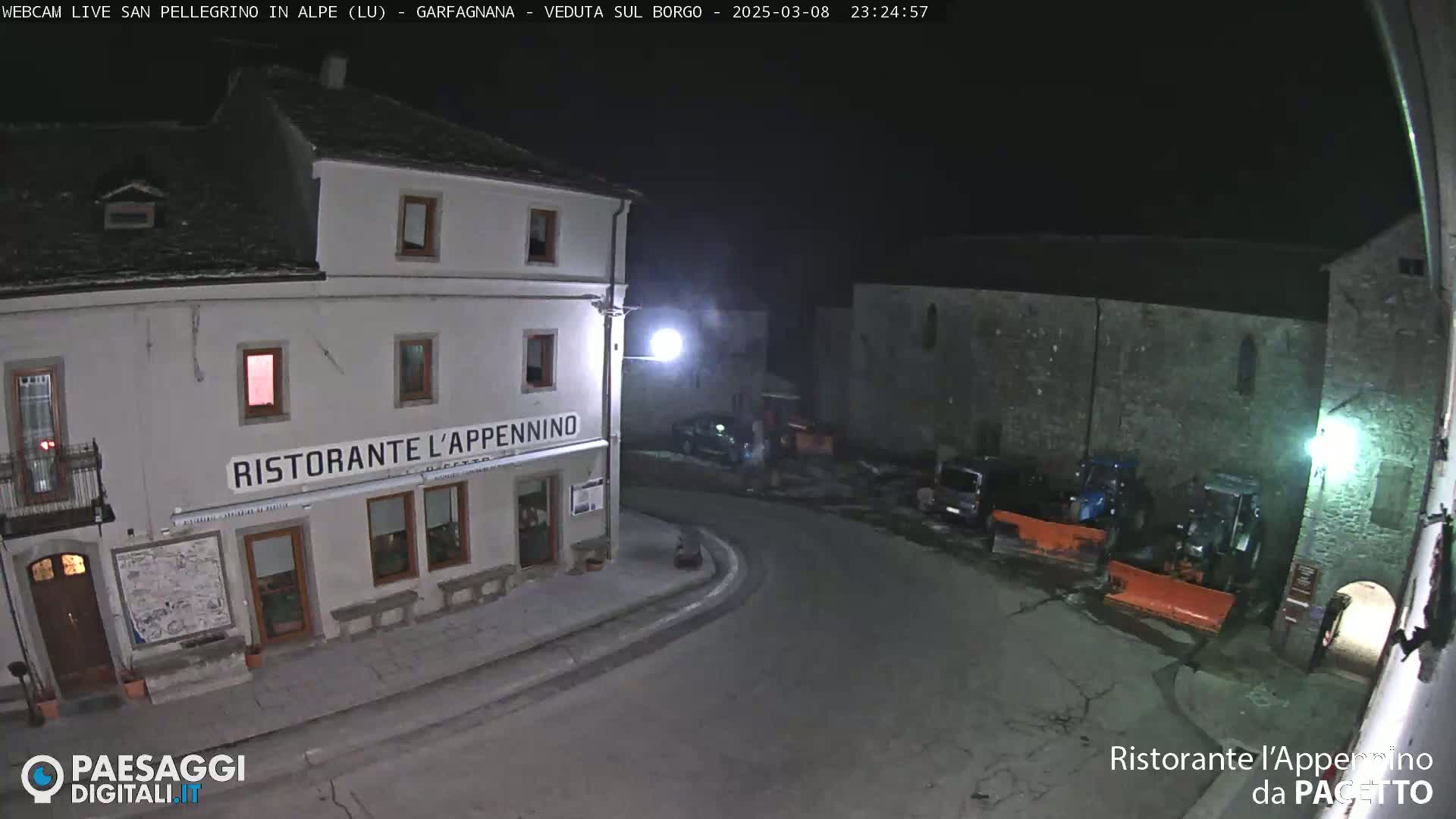 A nighttime, dimly lit town square features a restaurant, several parked vehicles, and snow-clearing equipment.