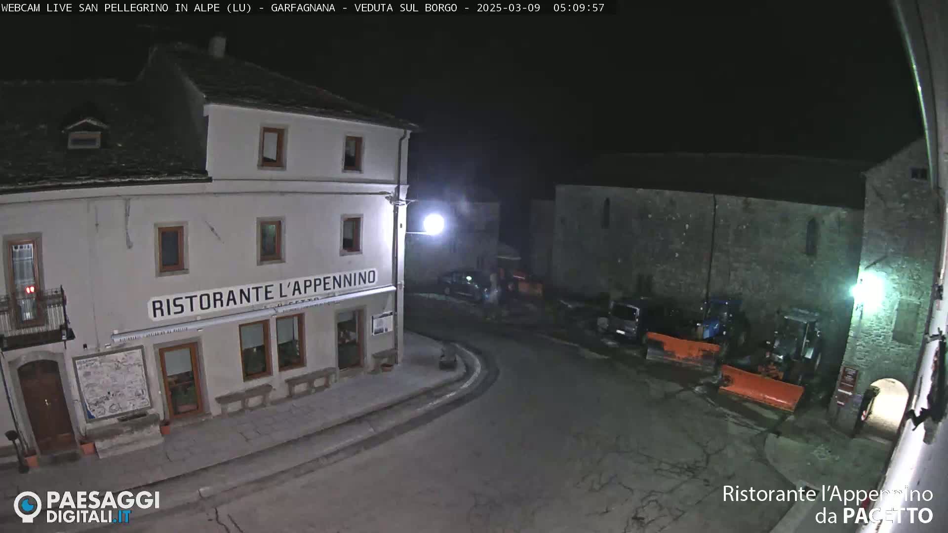 A nighttime, outdoor view of a town square shows a restaurant called "Ristorante L'Appennino" and several parked vehicles, including snowplows.
