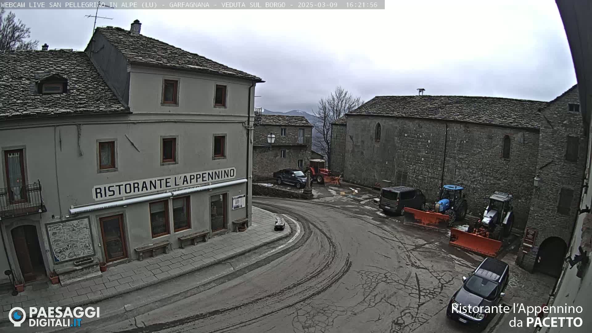 A town square with stone buildings, a restaurant, parked vehicles, and snow removal equipment under an overcast sky.