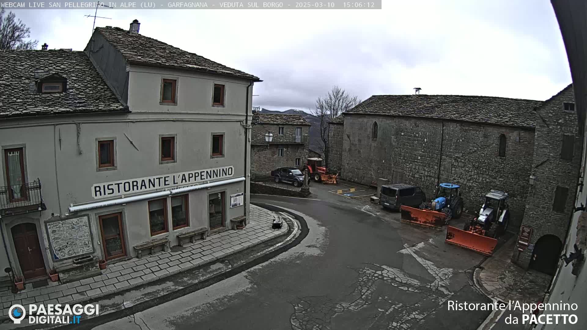 A village square with stone buildings, a restaurant, several vehicles including tractors and snow plows, under an overcast sky.