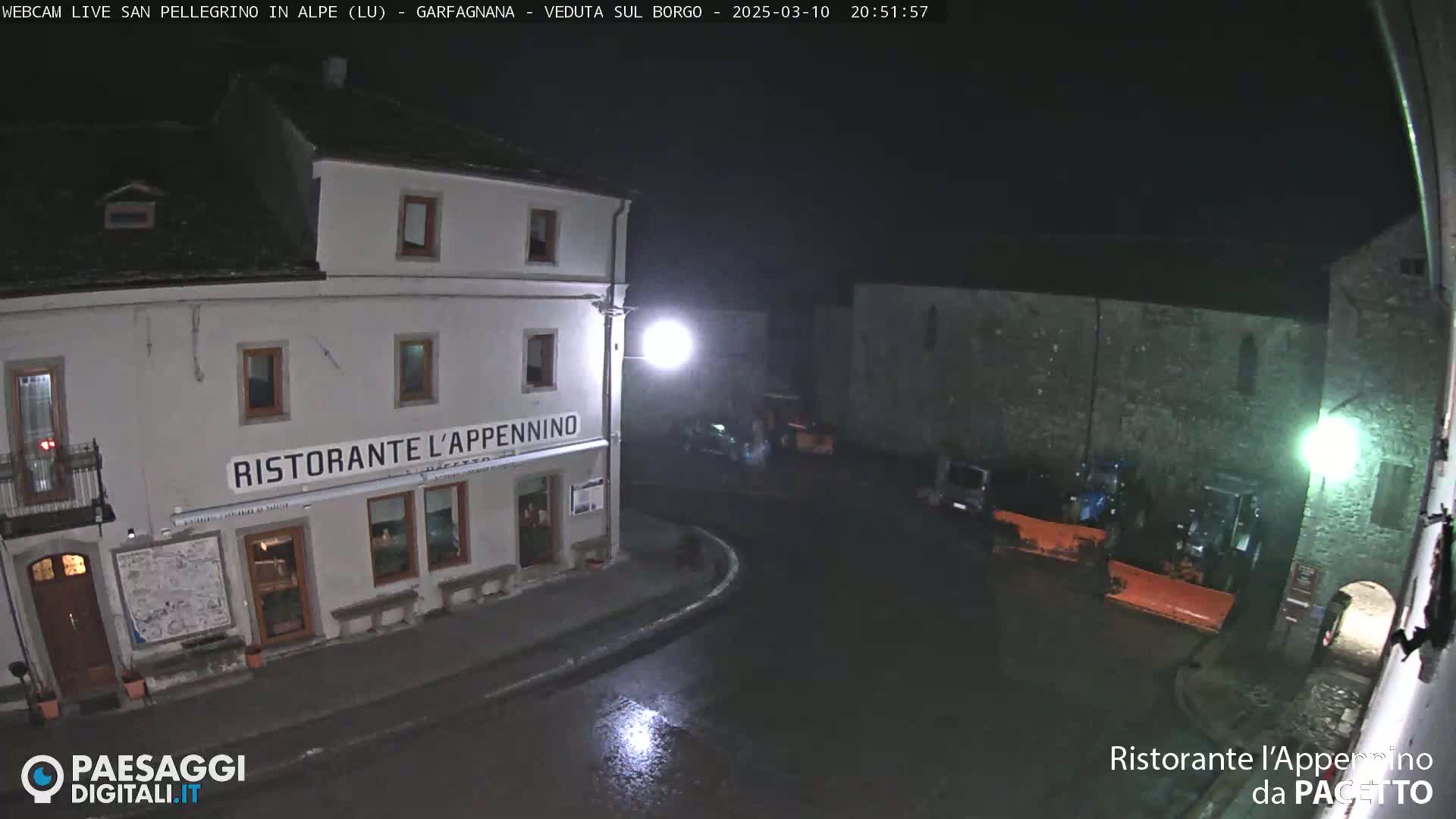 A nighttime view of a town square, wet from rain, showing a restaurant and snow plows parked near a stone building.