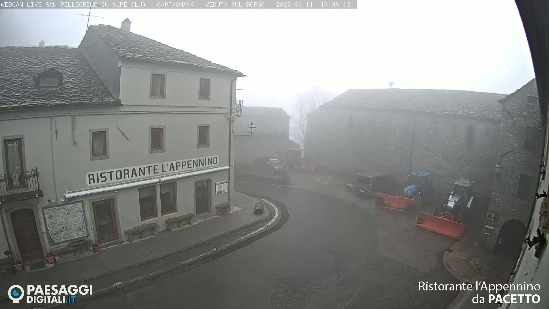 A foggy village square is seen, with a restaurant named "Ristorante L'Appennino" prominently featured near some parked vehicles and snow removal equipment.