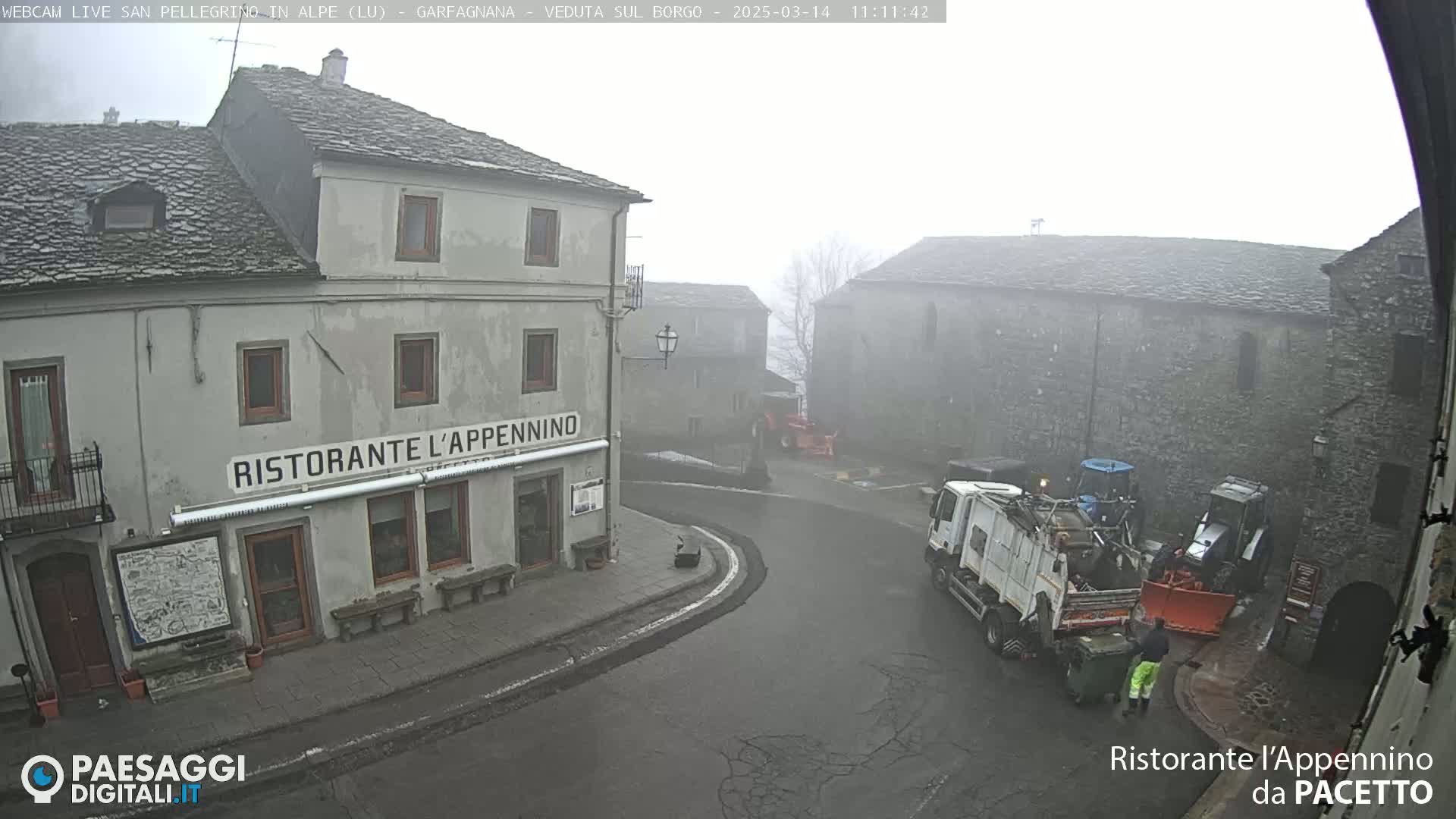 A foggy village square with stone buildings, a restaurant, and several pieces of machinery including a garbage truck and tractors.
