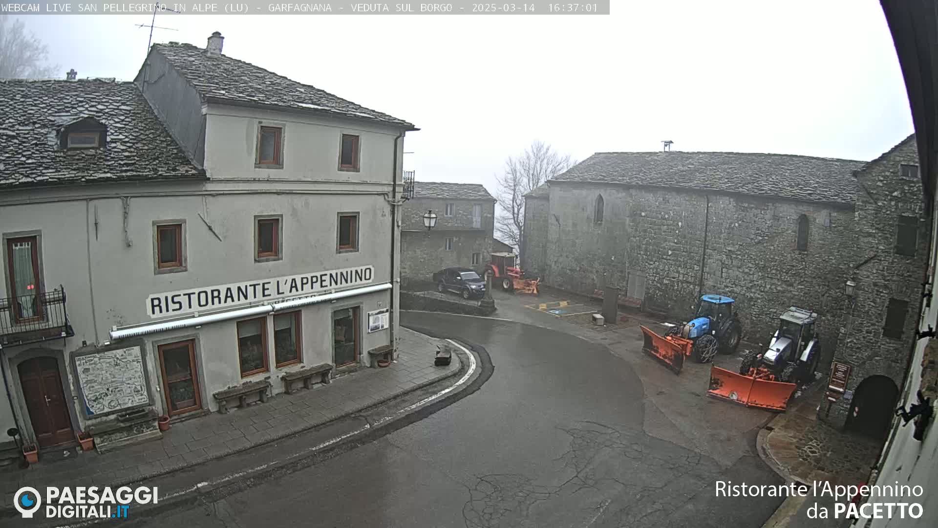 A foggy village square with stone buildings, a restaurant, parked vehicles, and snowplows.