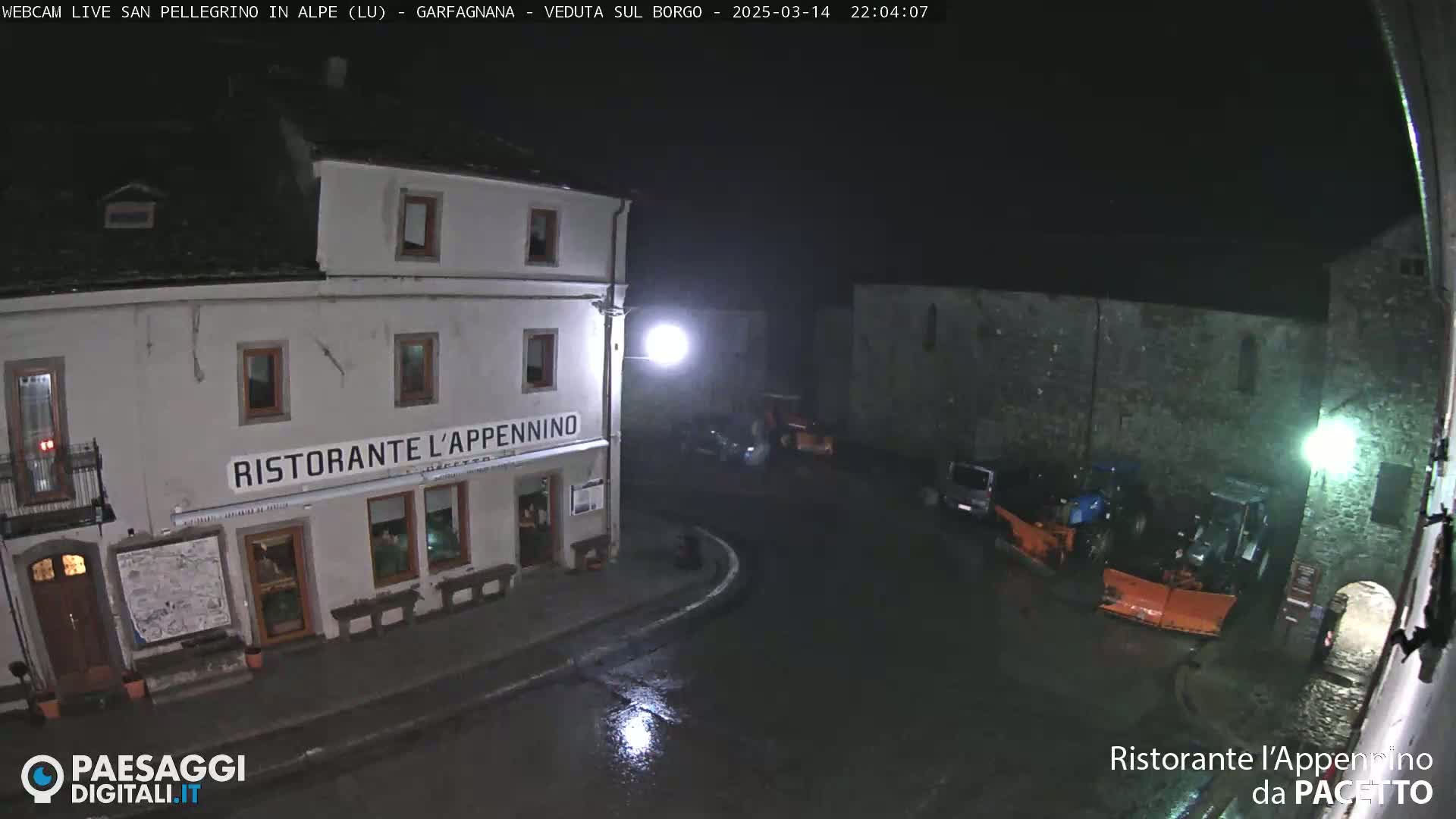 A nighttime view of a town square shows a restaurant, several parked vehicles, and snowplows under wet, dark conditions.