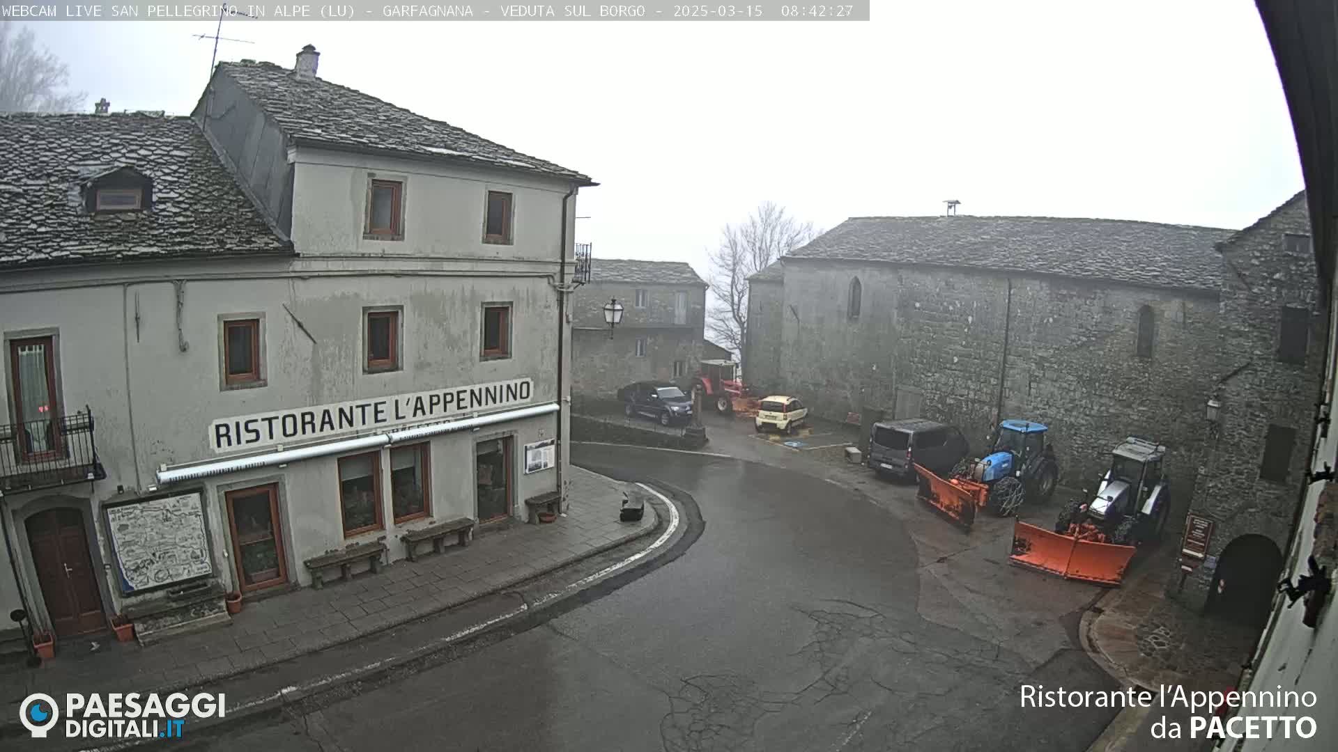 A foggy village square with several vehicles, including tractors and snowplows, parked near stone buildings.