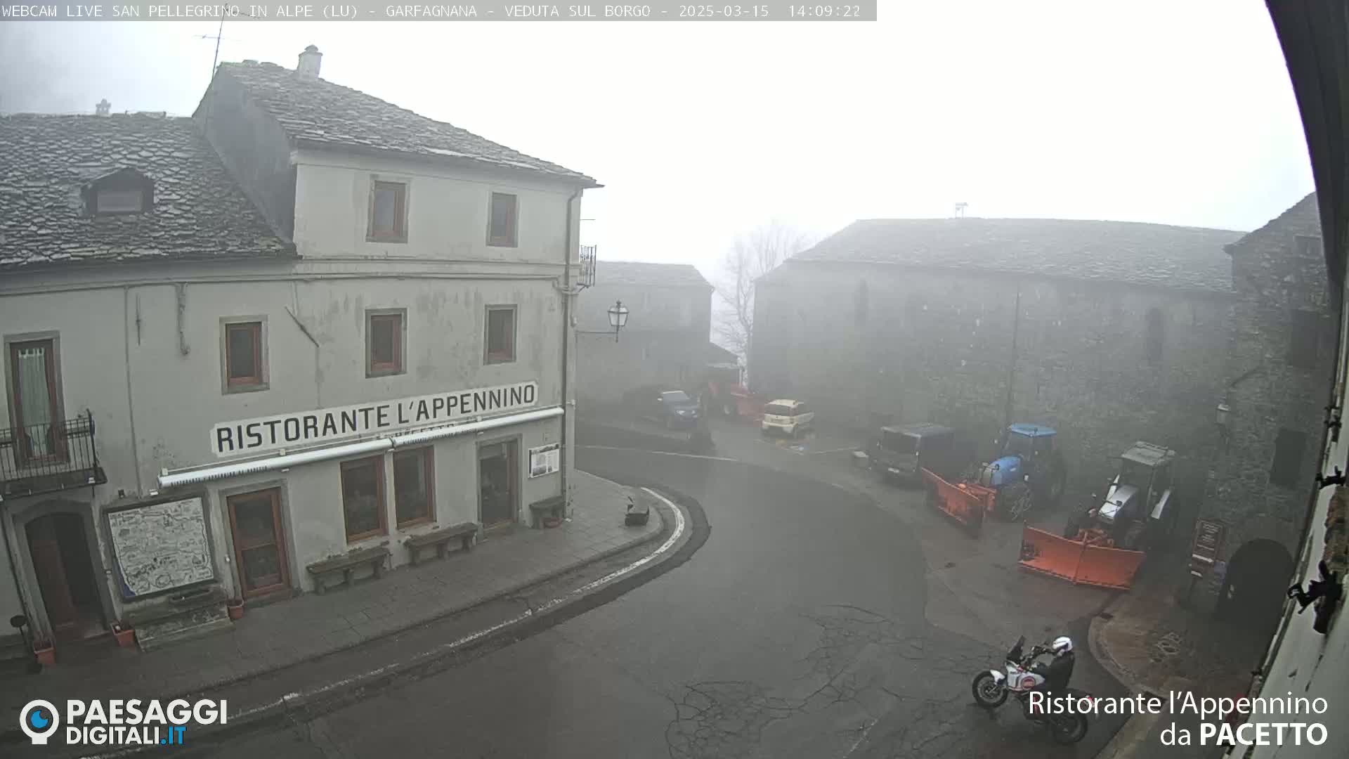 A foggy village square with stone buildings, a restaurant, several vehicles, and a motorcycle.