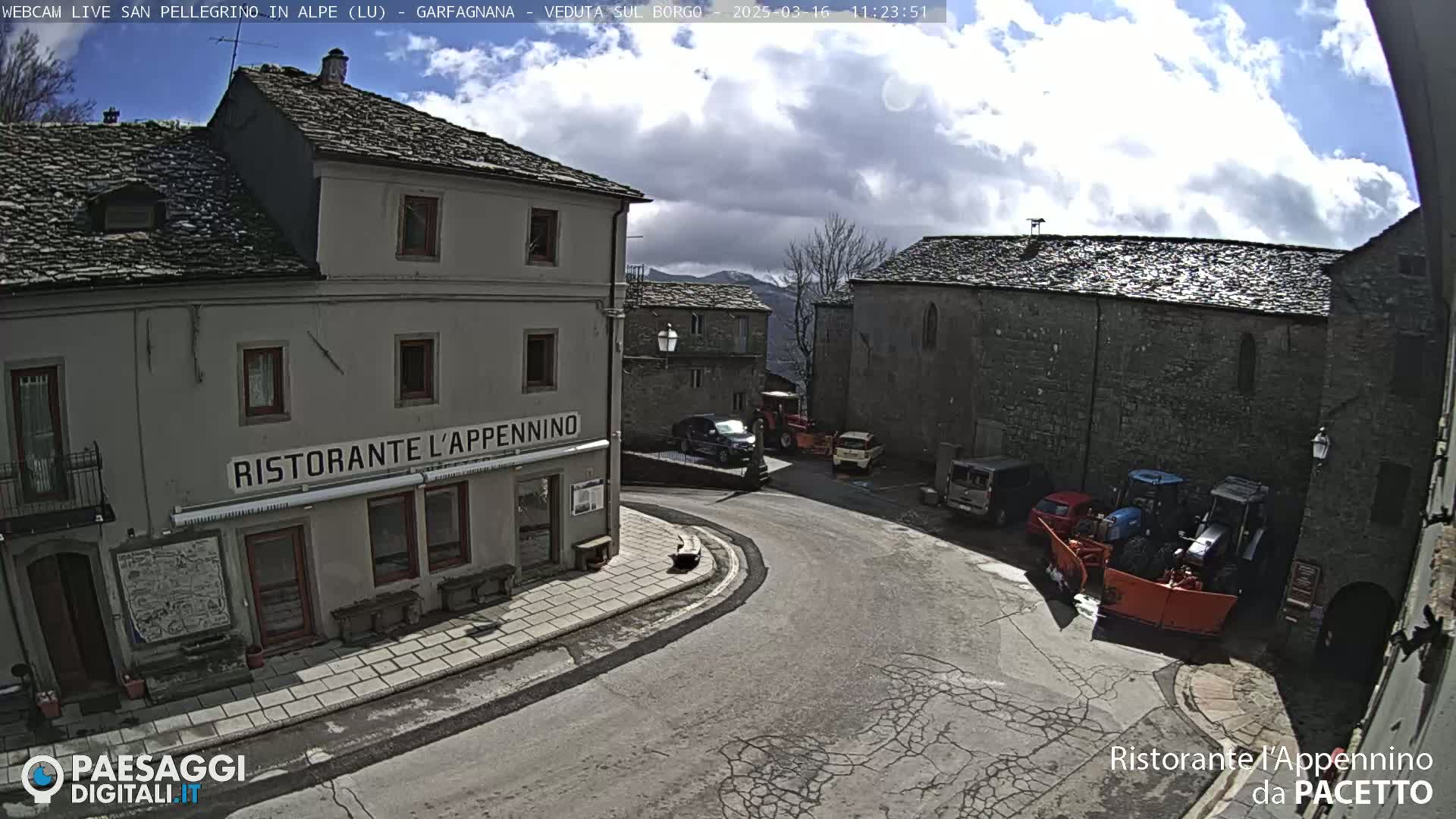A partly cloudy day reveals a village square with stone buildings, several cars, and snow removal equipment.