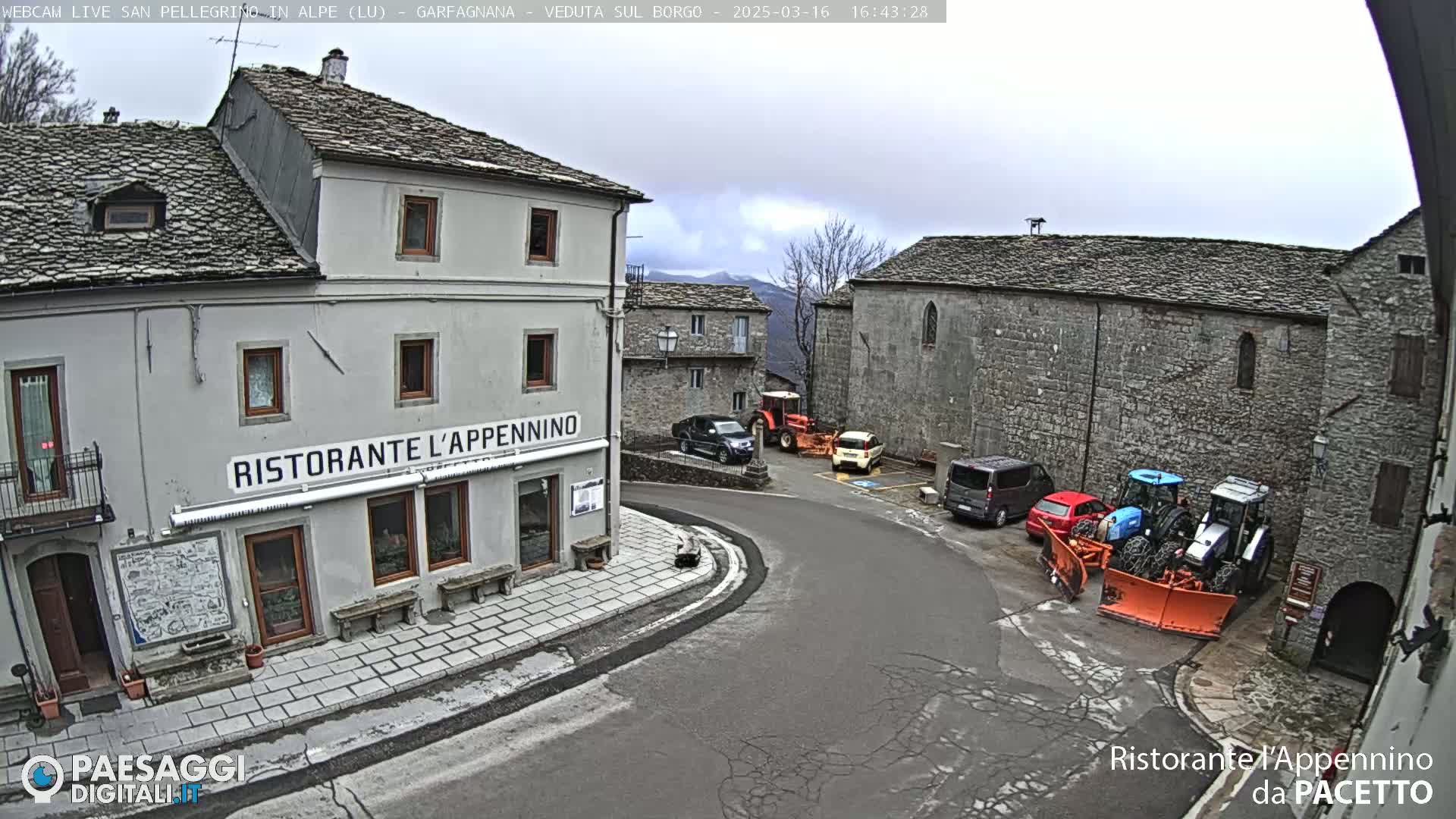 A stone village square on an overcast day, with several vehicles parked near a restaurant and some tractors with snow plows.