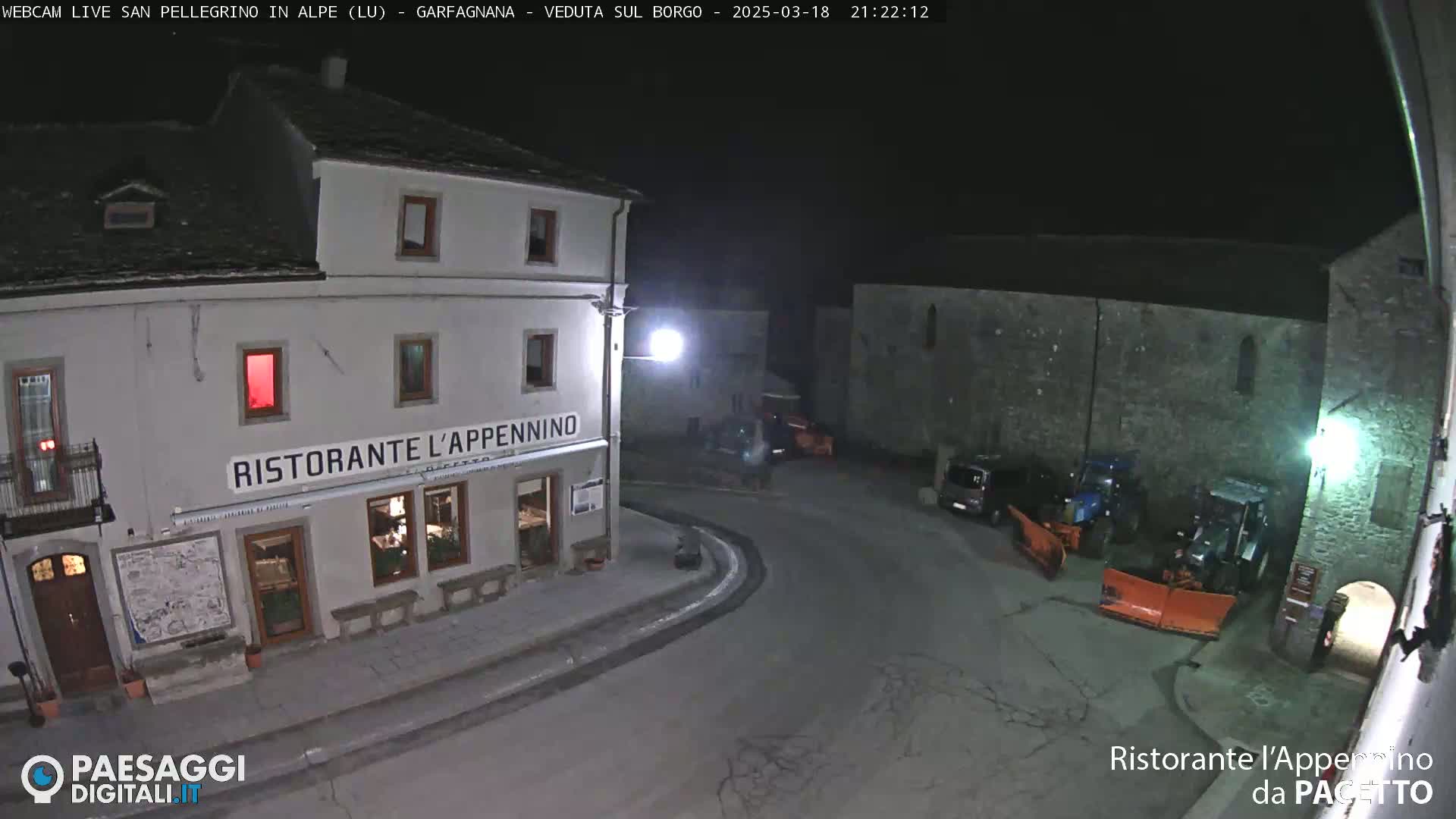 A nighttime view of a town square with a restaurant, several parked vehicles, and snowplows.
