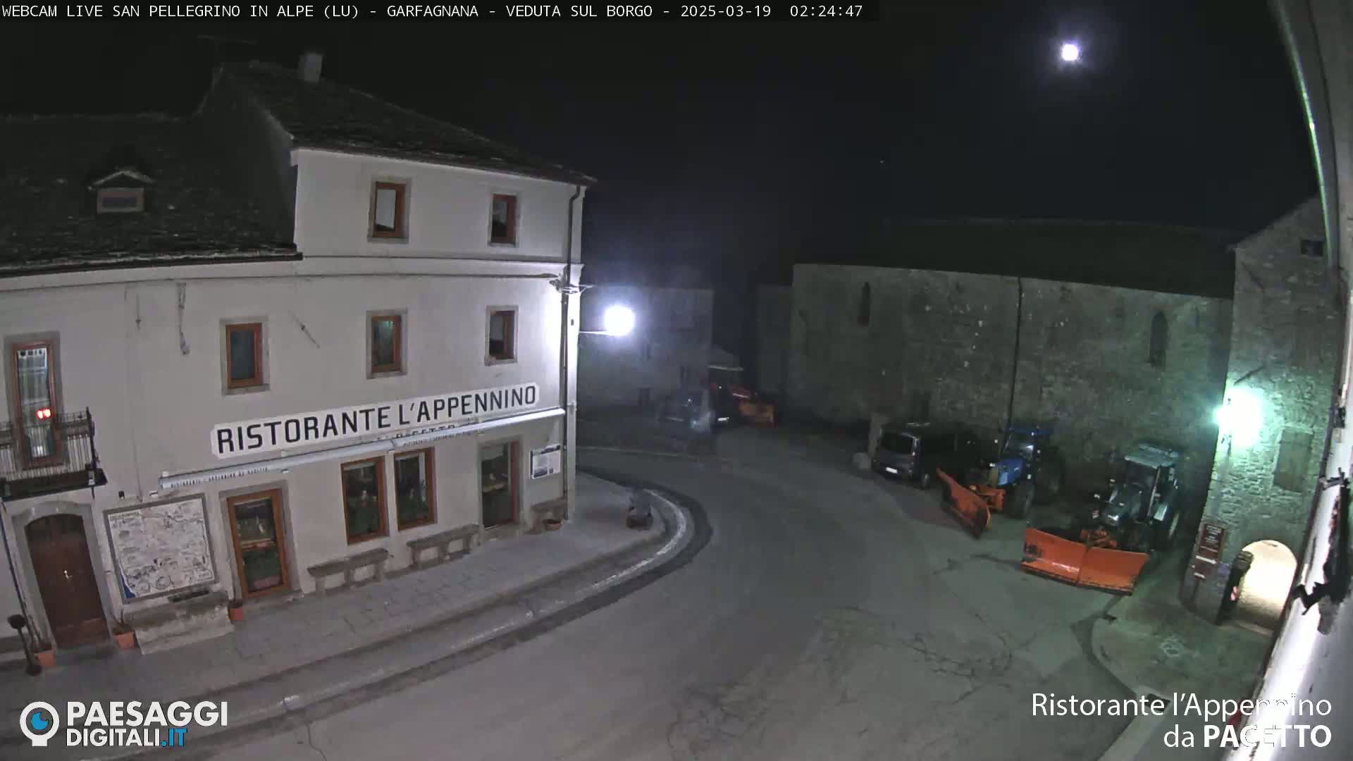 A nighttime view of a town square with a restaurant, some parked vehicles, and snowplows under a bright moon.
