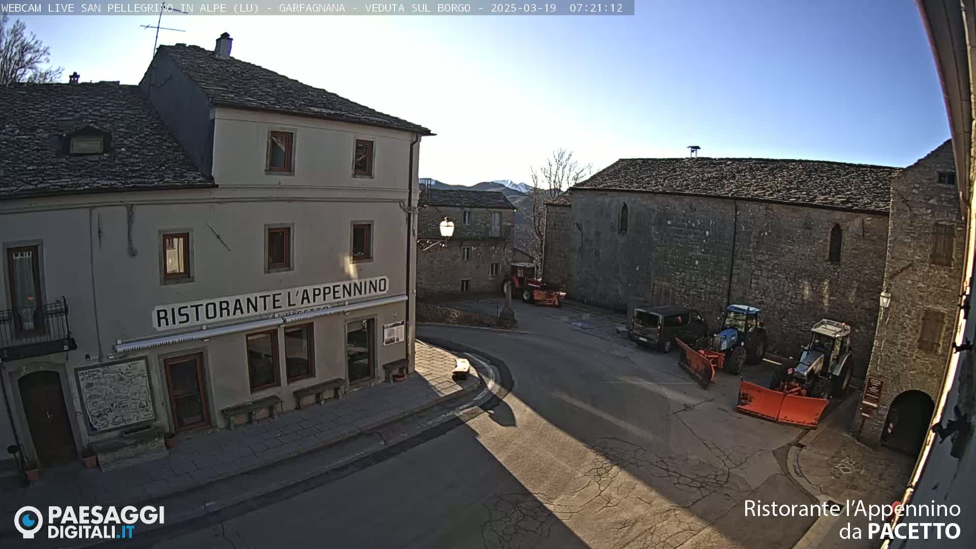 A sunny view of a town square with stone buildings, several parked tractors, and a restaurant.