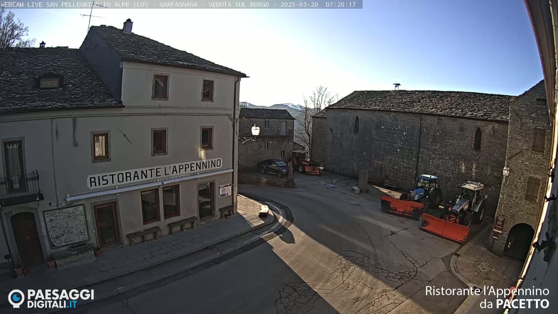 A sunny, high-angle view of a town square with stone buildings, a restaurant, and two tractors with snow plows.