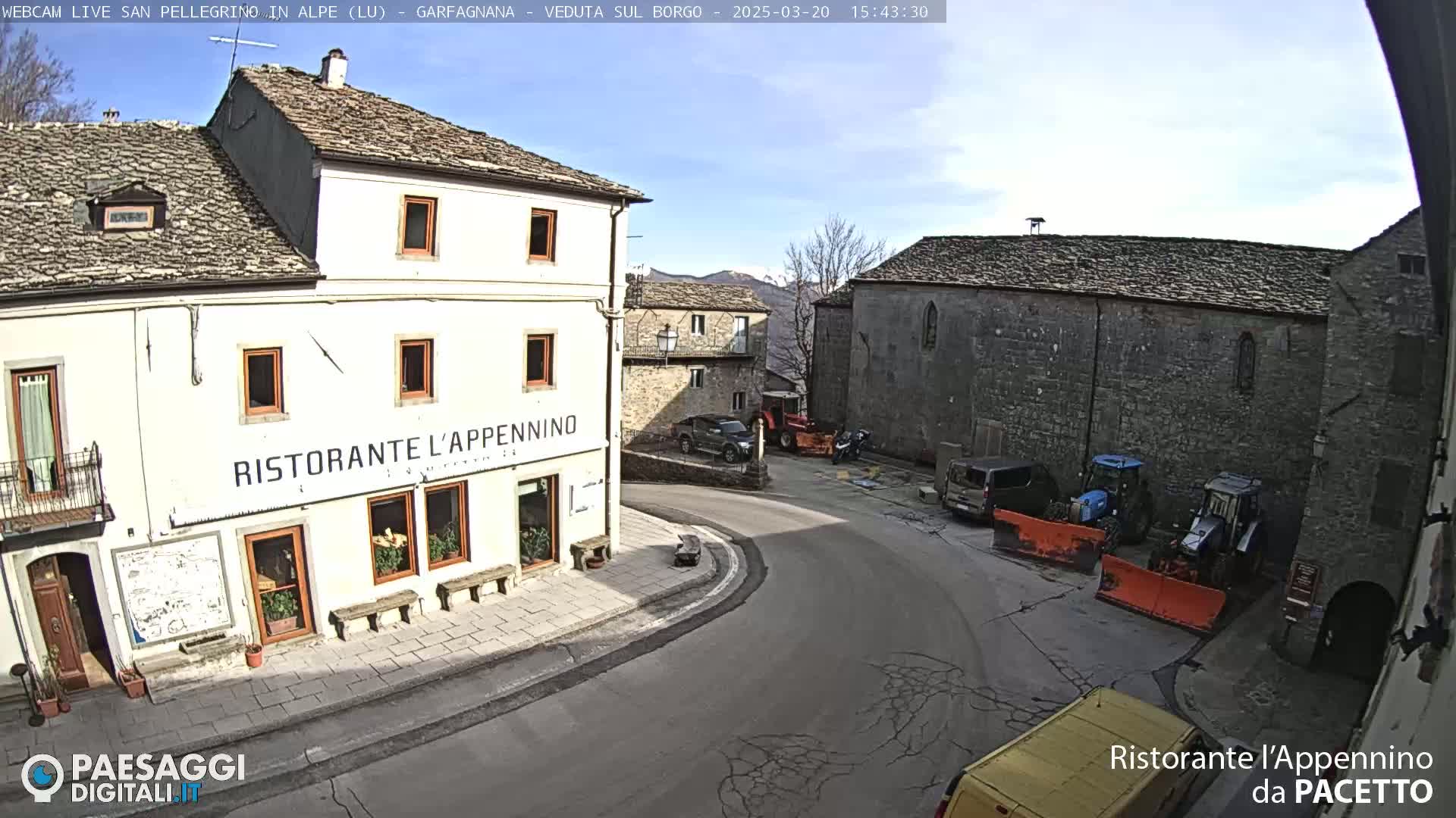 A sunny day reveals a quaint European village square with stone buildings, a restaurant, several vehicles, and some snow-removal equipment.