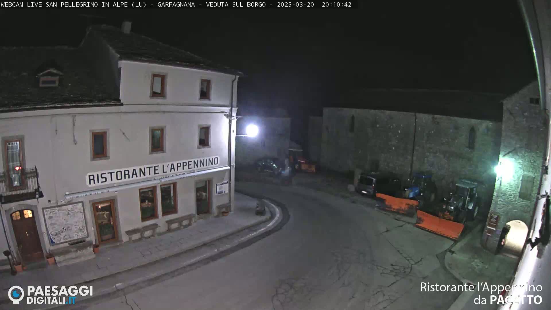 A nighttime view of a town square with a restaurant, several parked vehicles, and snow removal equipment under a dark sky.