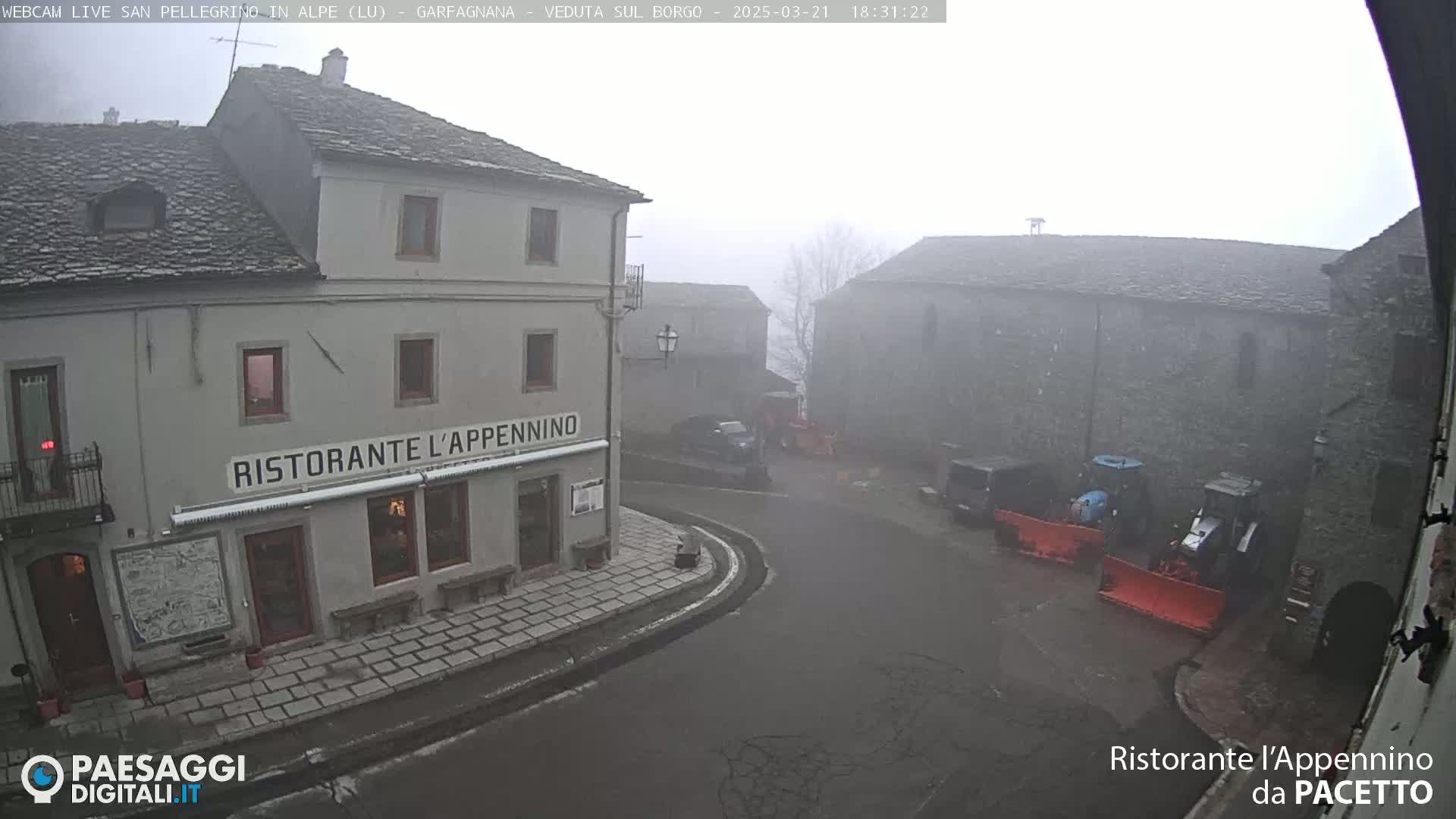 A foggy village square with a restaurant and several vehicles, including snowplows, is visible.