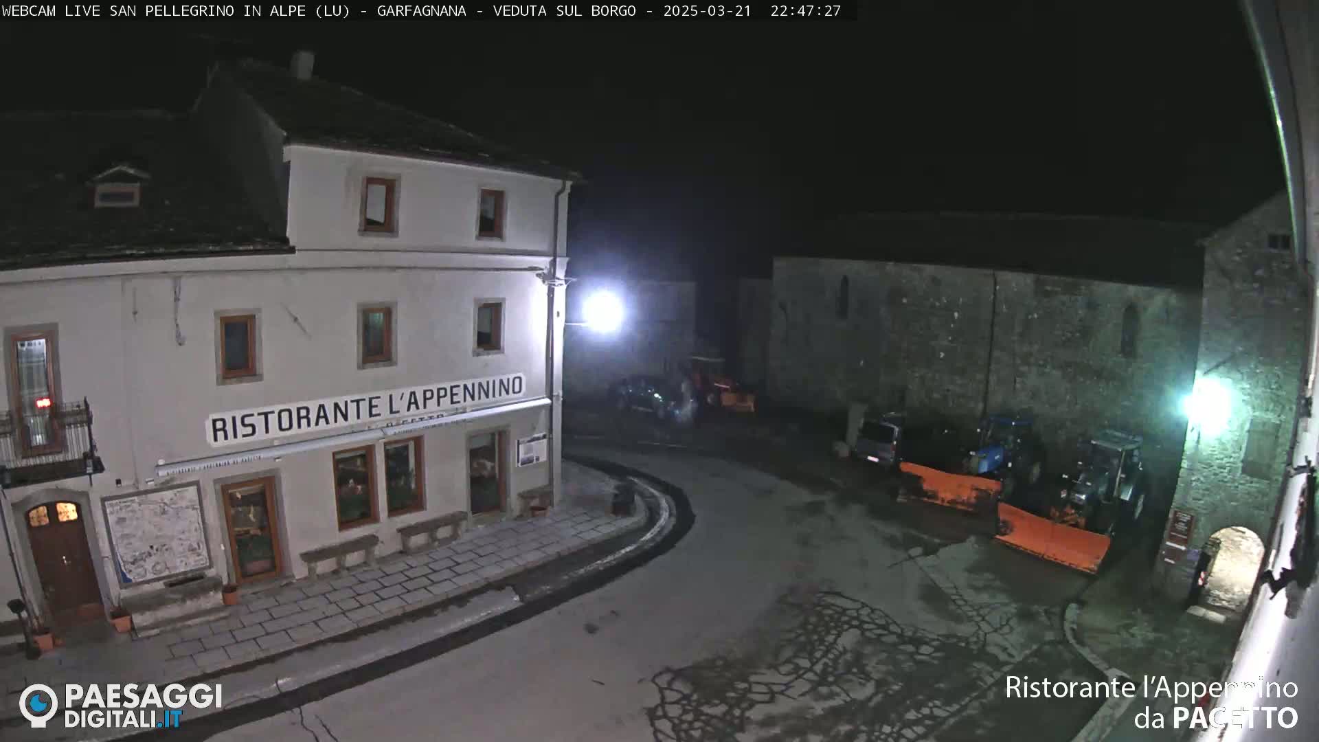 A nighttime view of a town square with a restaurant, some parked cars, and snow-removal equipment under dark, clear skies.