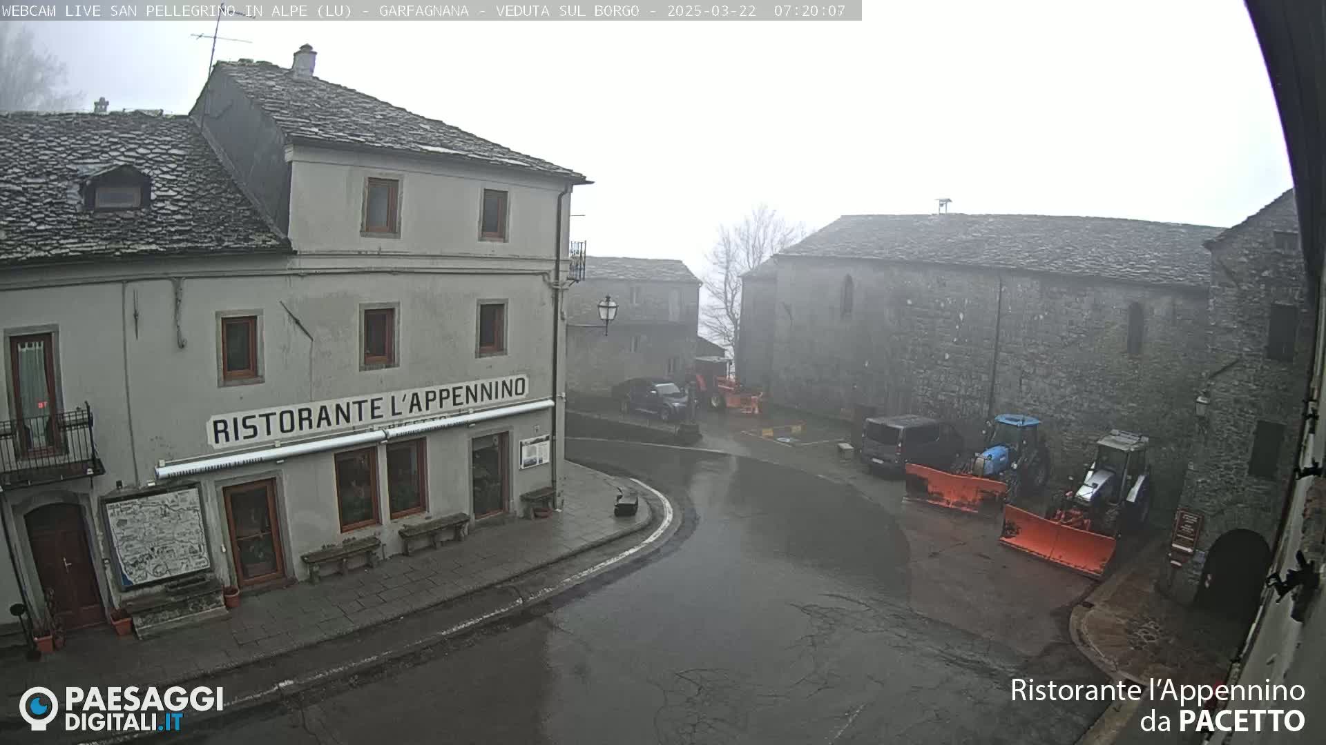 A wet, foggy village square with several stone buildings, a restaurant, and snowplows parked near a street corner.